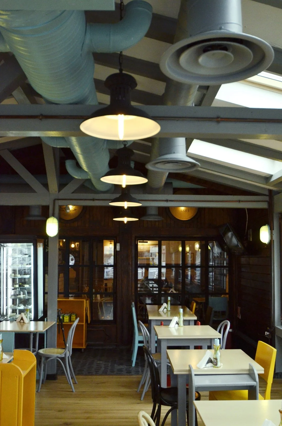 Interior view of a cozy restaurant with white tables, colorful chairs, and hanging lights, featuring visible ductwork on the ceiling.