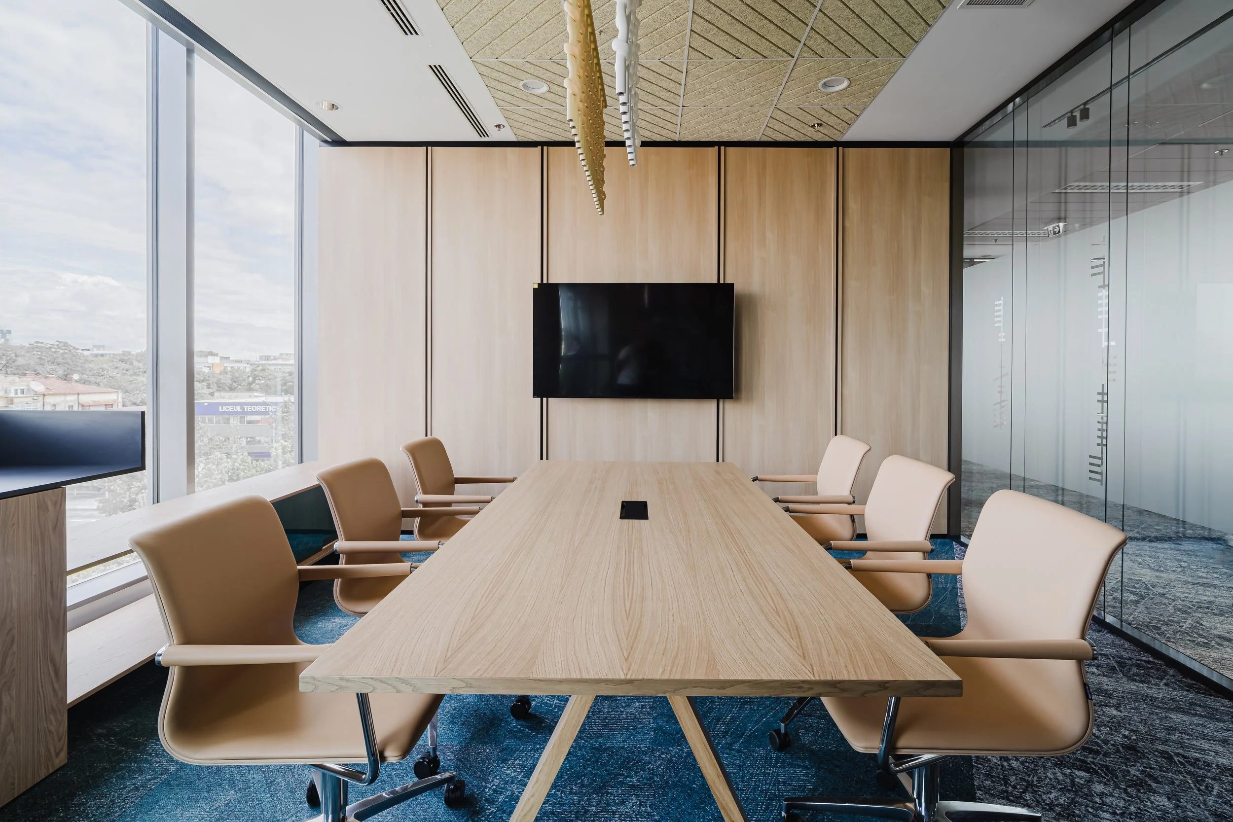 Modern conference room with a long wooden table, eight beige chairs, a large flat-screen TV on the wall, and floor-to-ceiling windows showing a cloudy sky and cityscape.