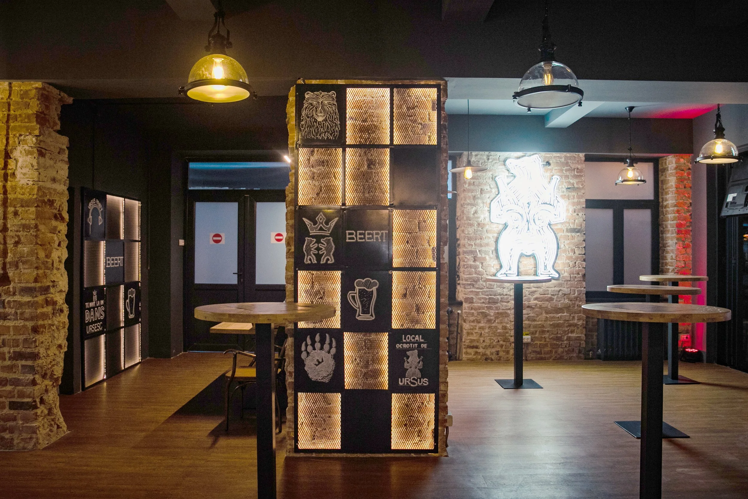 Interior of a bar or pub with exposed brick walls, wooden floors, decorative lighting fixtures, and illuminated neon or sign art, including a lion or bear head and beer-related signs.