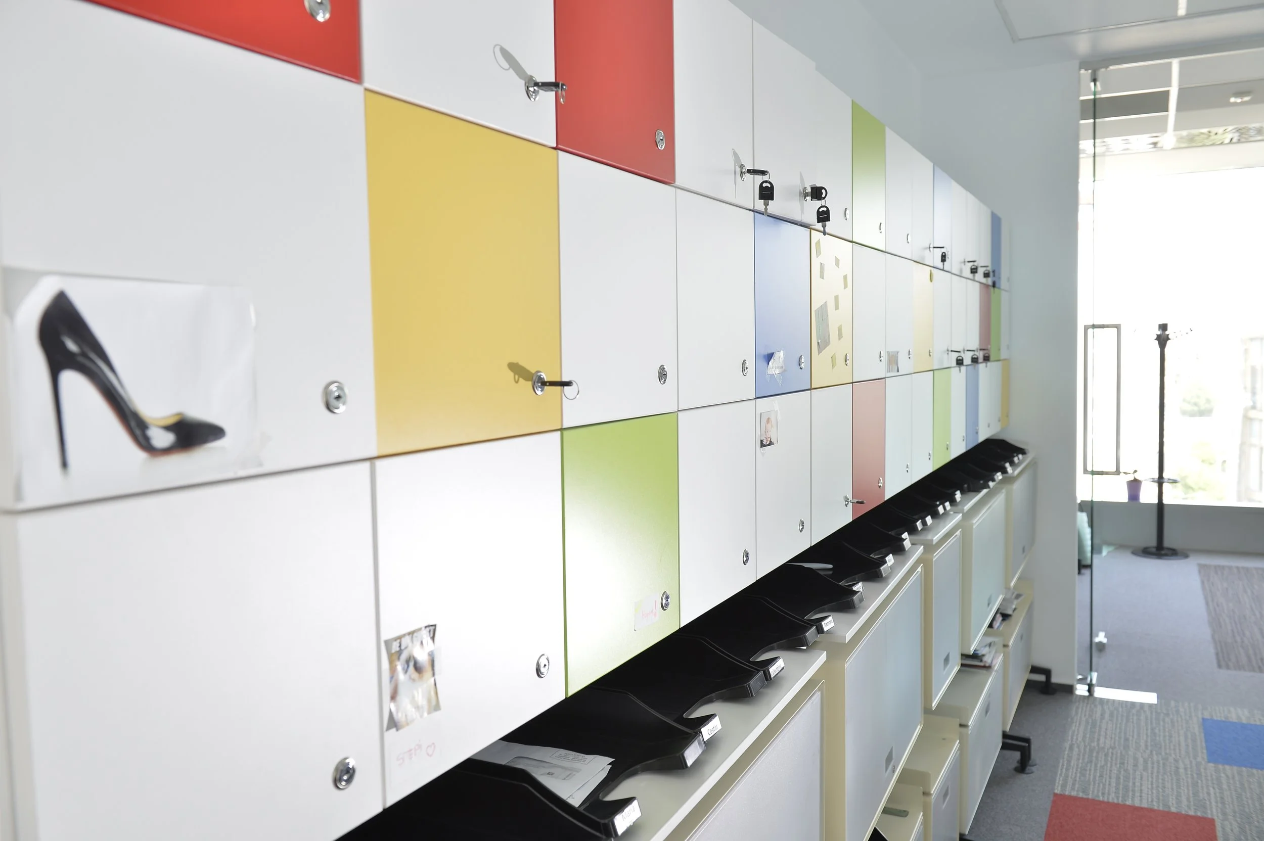 Colorful lockers with locks and a few photos and artwork, in an office or lobby area. Some lockers are open, showing black trays inside.