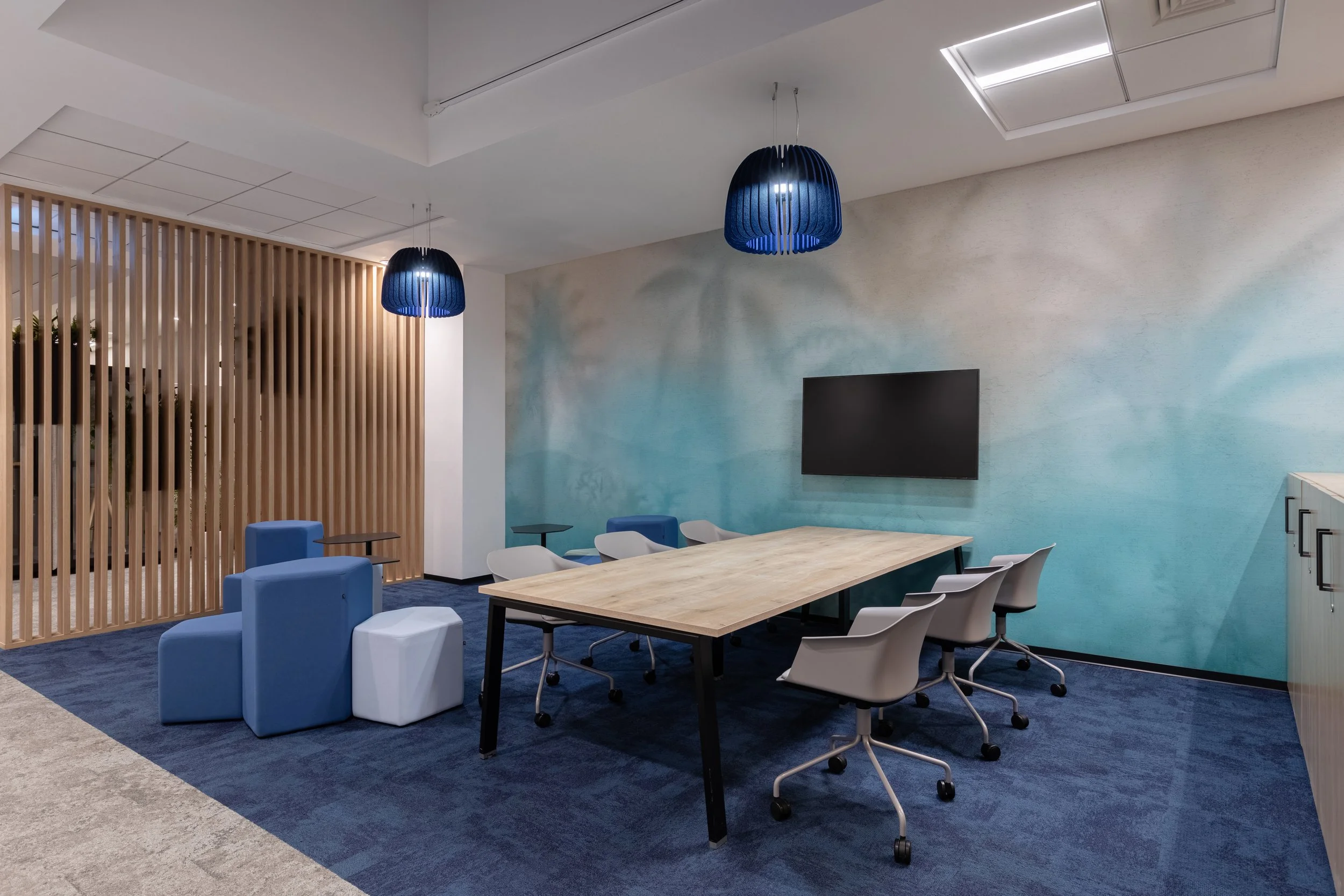 Modern conference room with a large wooden table, six gray chairs on wheels, blue and white ottomans, two blue hanging pendant lights, a wall-mounted flat-screen TV, a wooden slat room divider, and a textured blue and beige wall.