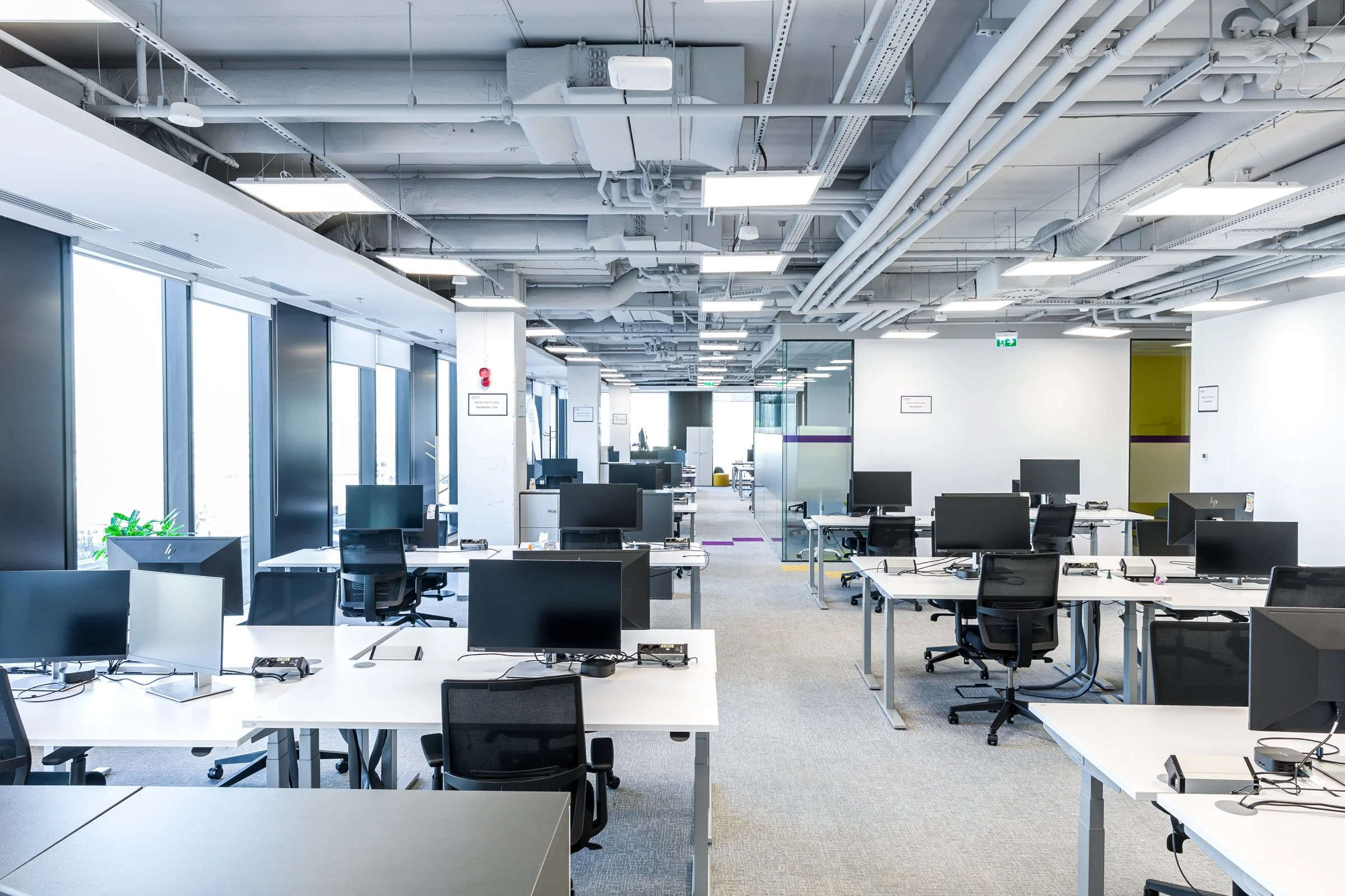 Empty modern office with multiple desks and computers, large windows, and exposed ceiling pipes.