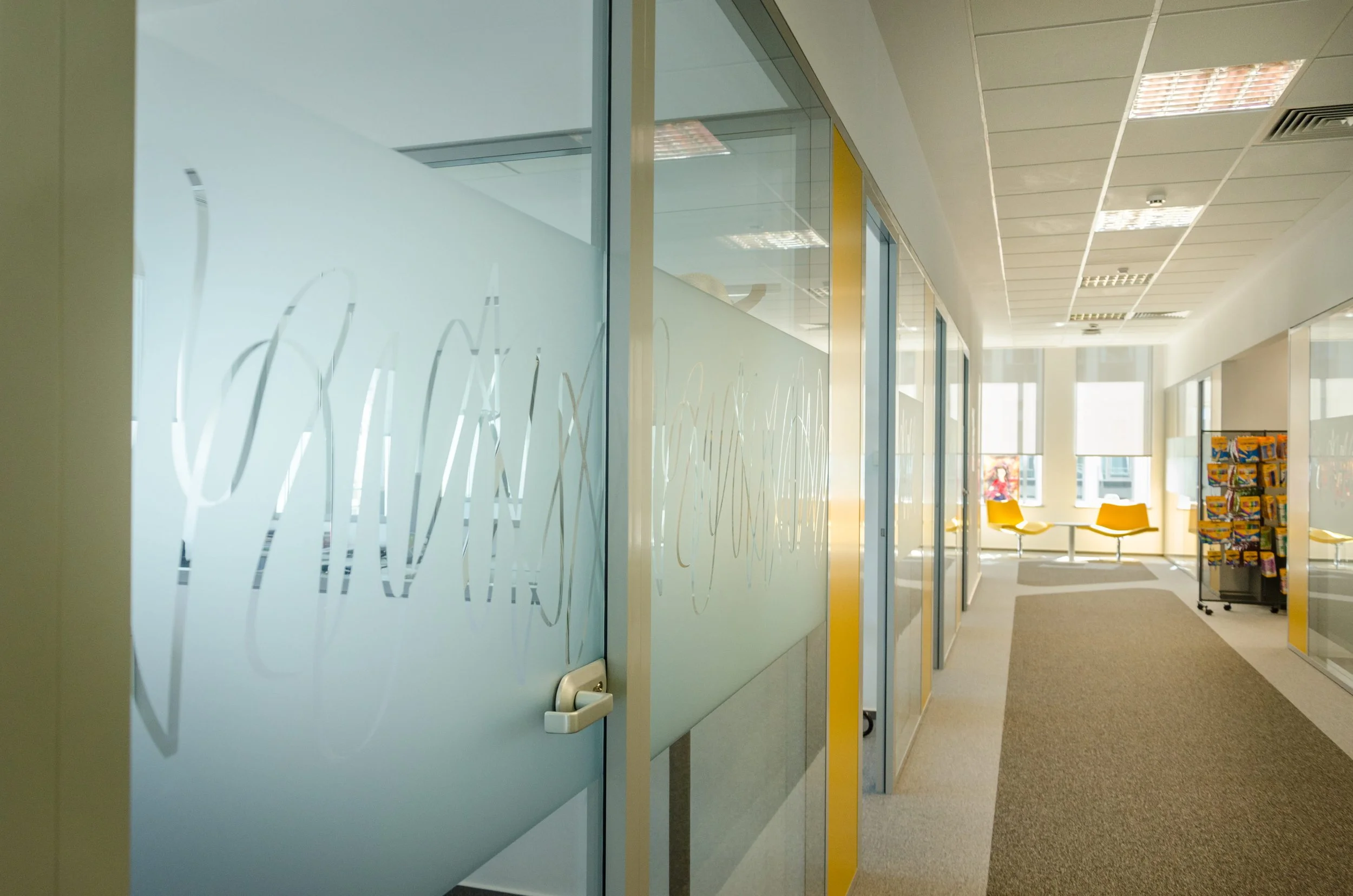 Empty modern office hallway with glass partition walls, yellow chairs at the end, a magazine rack on the right, and windows letting in natural light.