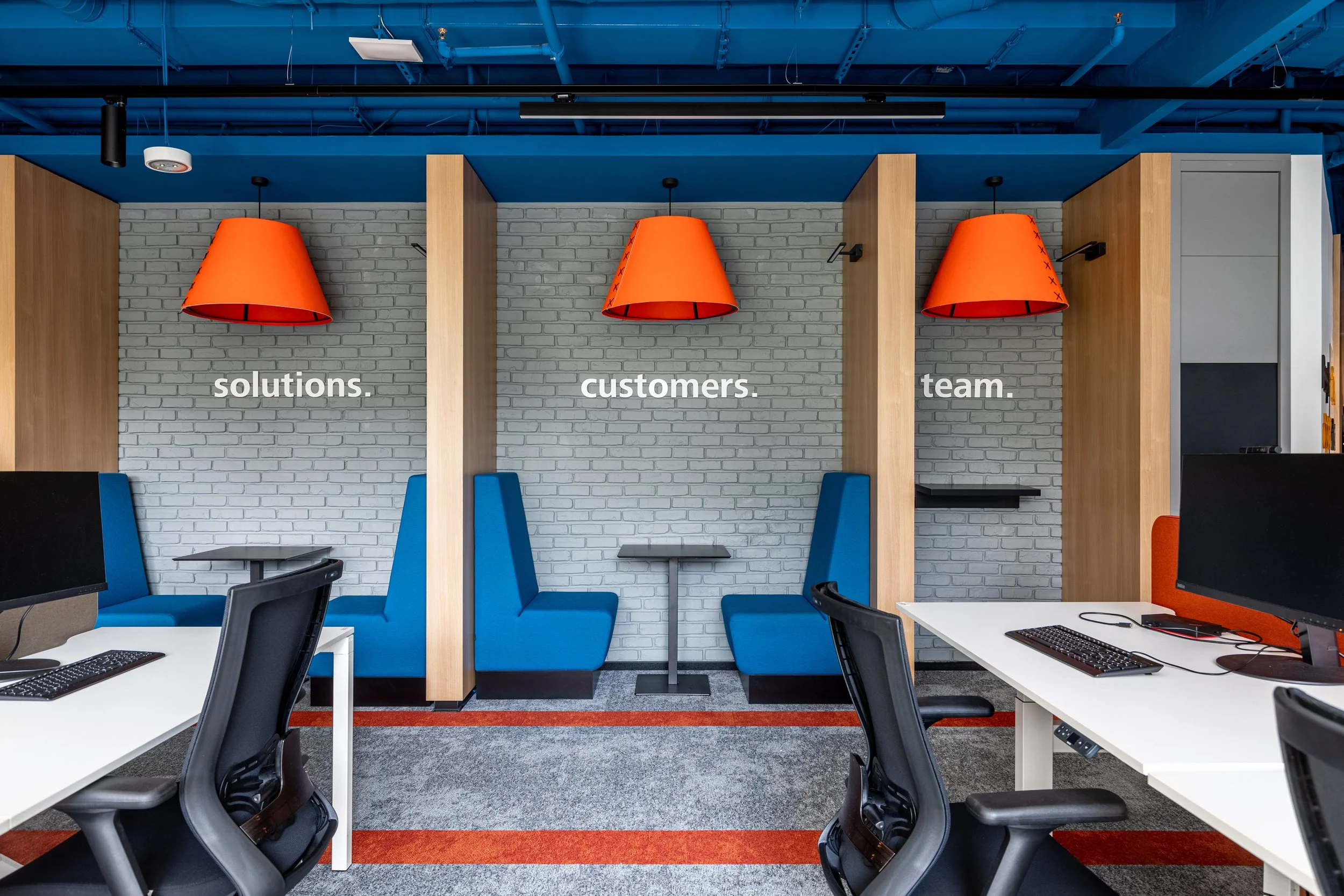 Modern office space with white desks, black chairs, blue booth seating, and orange pendant lights. Words on the wall say 'solutions.', 'customers.', and 'team.'.