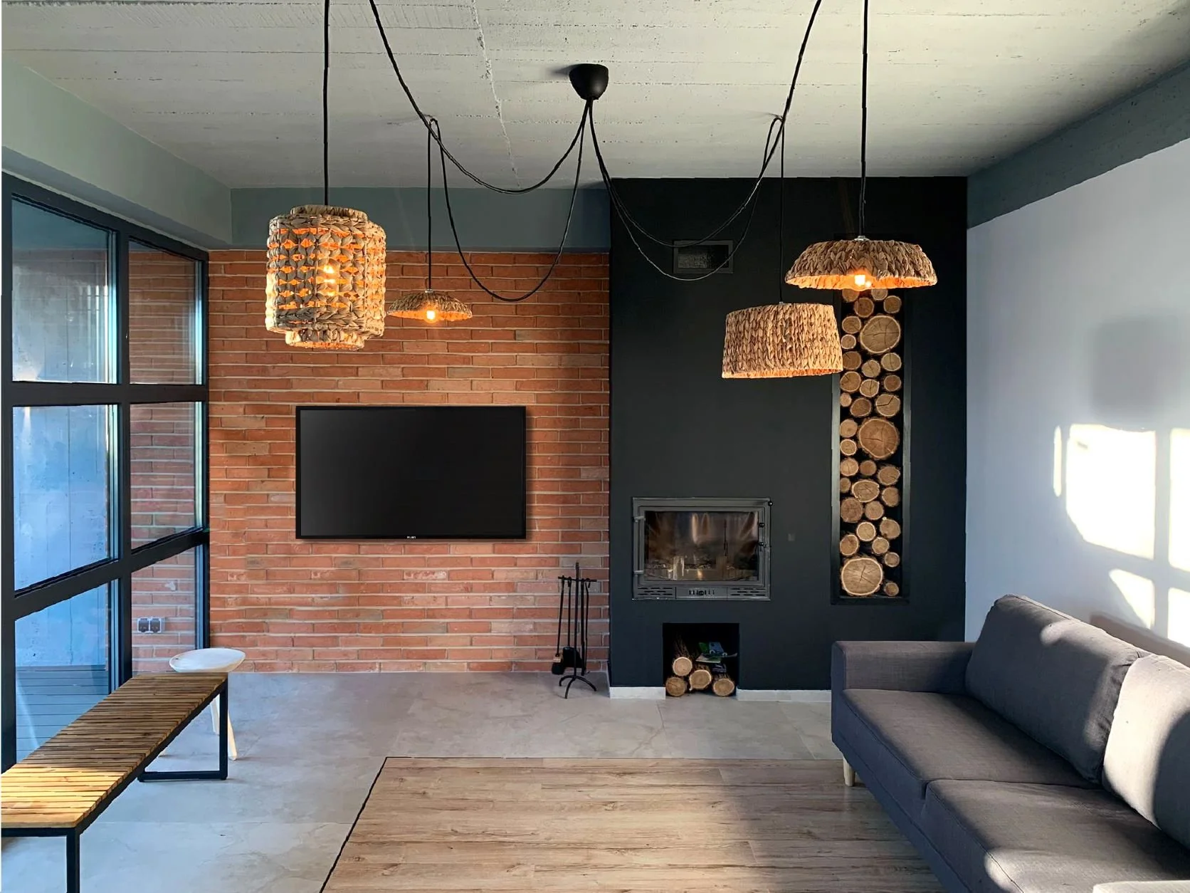 Living room with brick wall, mounted flat-screen TV, black fireplace with stacked firewood, gray sofa, wooden bench, pendant lights with woven shades, large window, and concrete flooring.