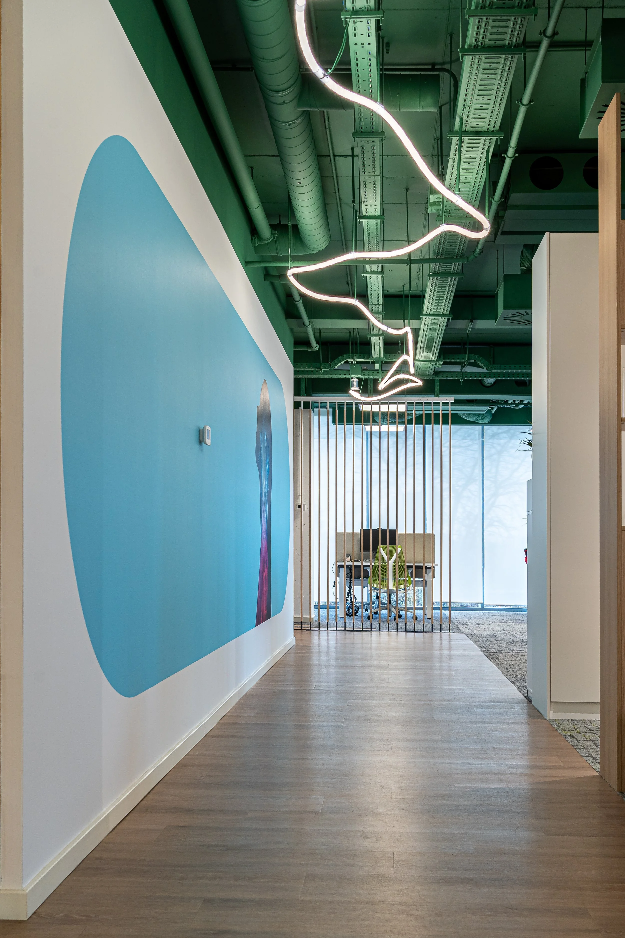 Modern office hallway with green ceiling and exposed ductwork, featuring a neon light sculpture hanging from the ceiling, a blue rounded abstract mural on the wall, and a small office with a desk and chairs in the background near large window.