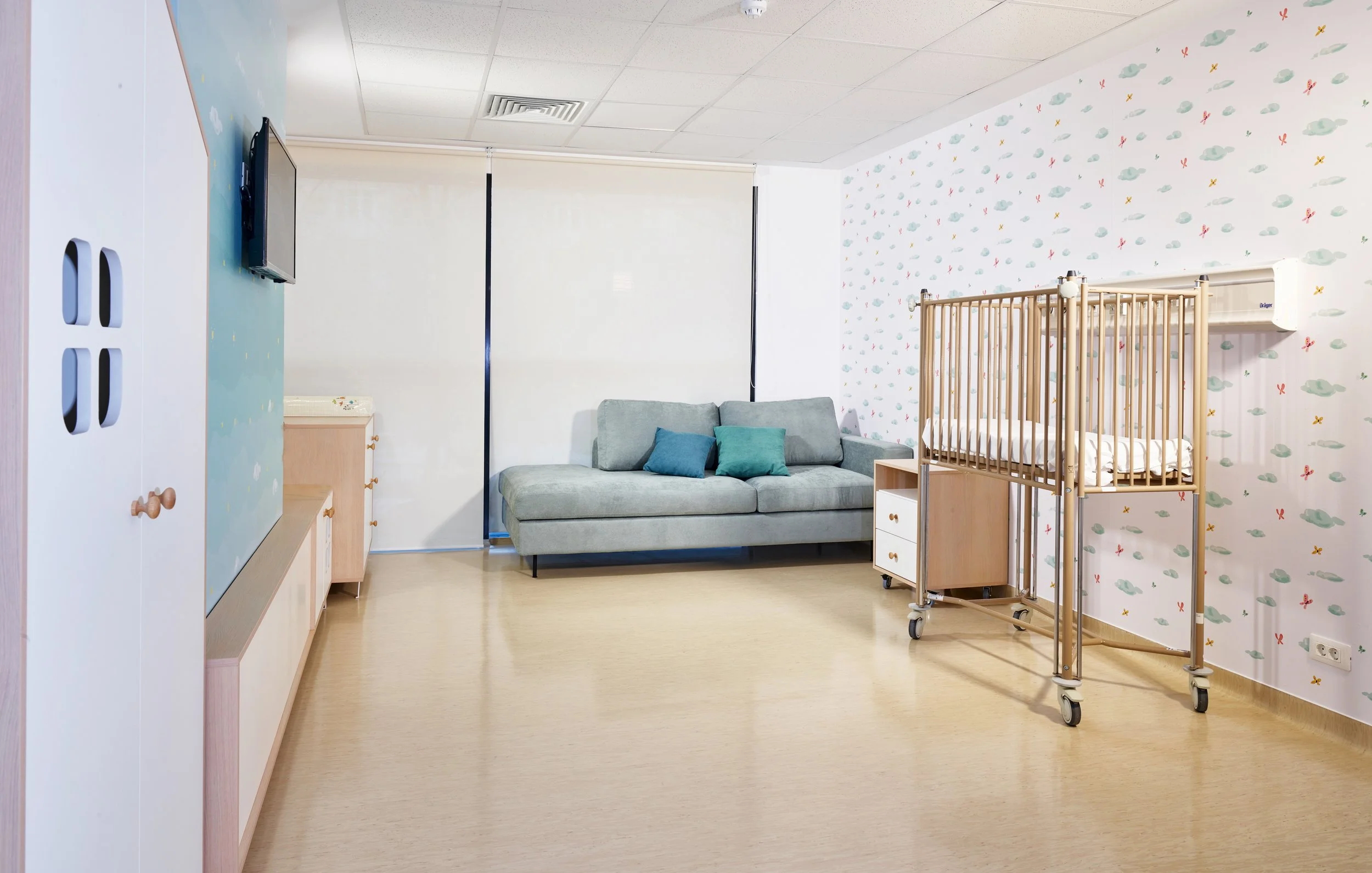 A nursery room with a gray sofa, light wood furniture, a crib with a mobile, a wall-mounted TV, and patterned wallpaper with clouds and butterflies.