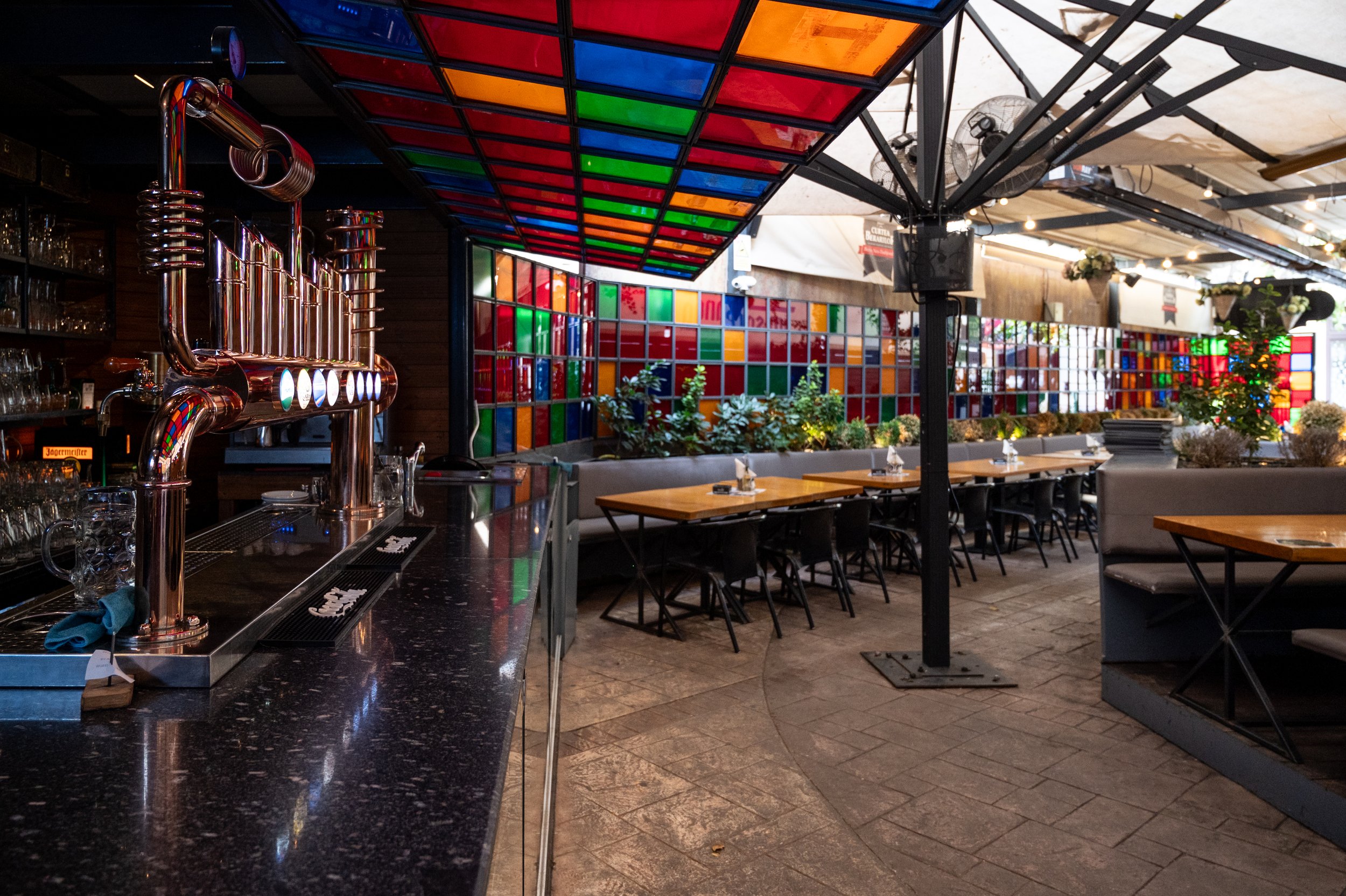 Inside a vibrant restaurant or bar with a colorful stained glass ceiling and wall decor, tables and chairs arranged for dining, a bar counter with beer taps, and decorative plants along the wall.