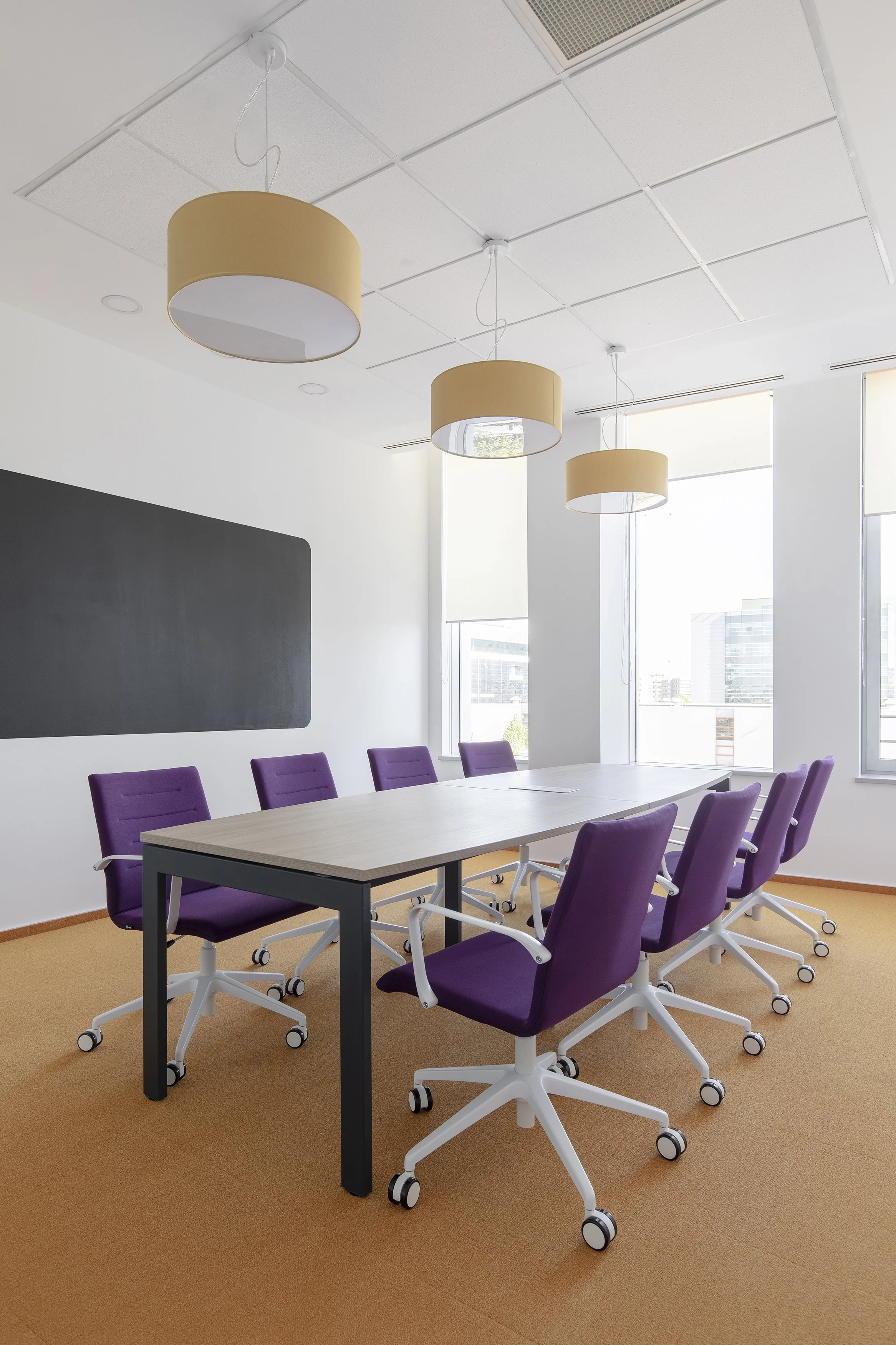 Empty conference room with six purple office chairs around a light wood table, three beige pendant lights hanging from a white ceiling, large windows letting in natural light, and a blackboard on the wall.