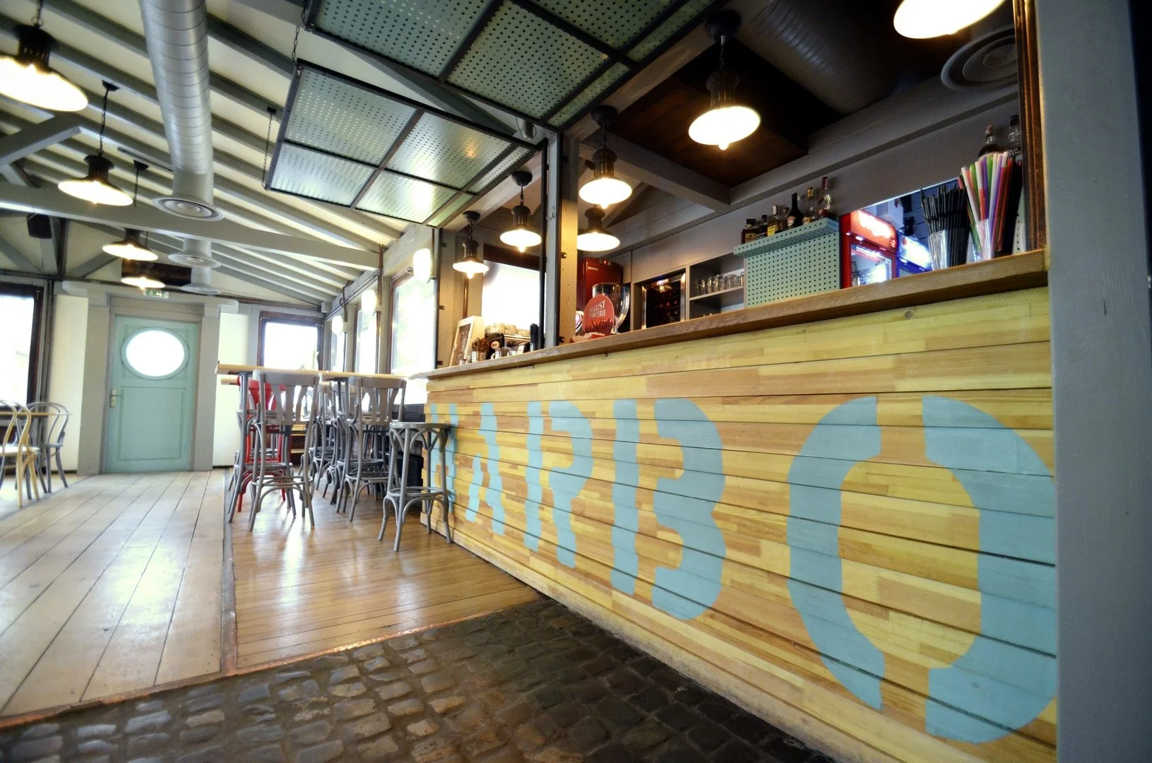 Interior view of a cafe or restaurant with a wooden counter and chairs, colorful drinks and bar equipment behind the counter, and a door with an oval window. The space has a mix of natural and artificial lighting.