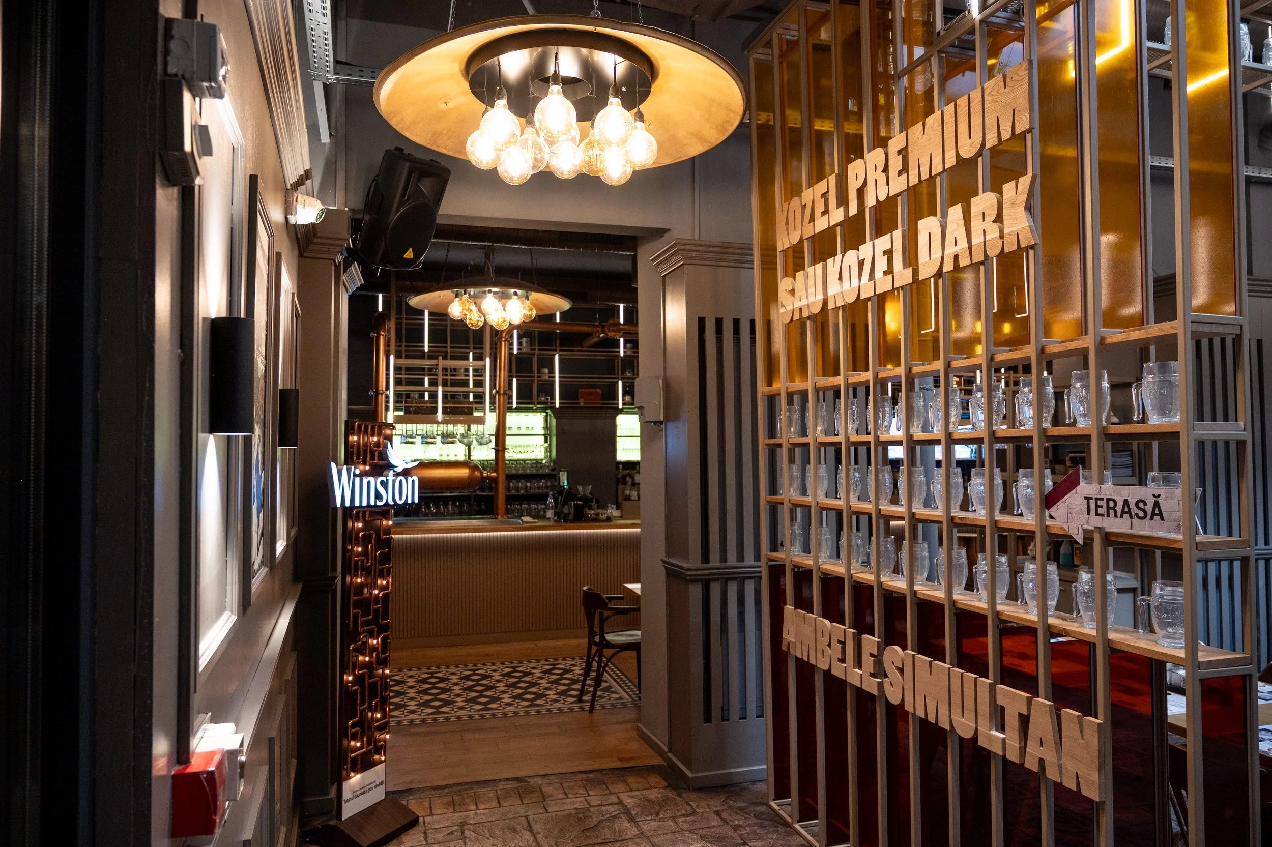 Interior of a stylish bar or restaurant with warm lighting and modern decor, featuring a wooden lattice partition with glass jars and hanging signs.