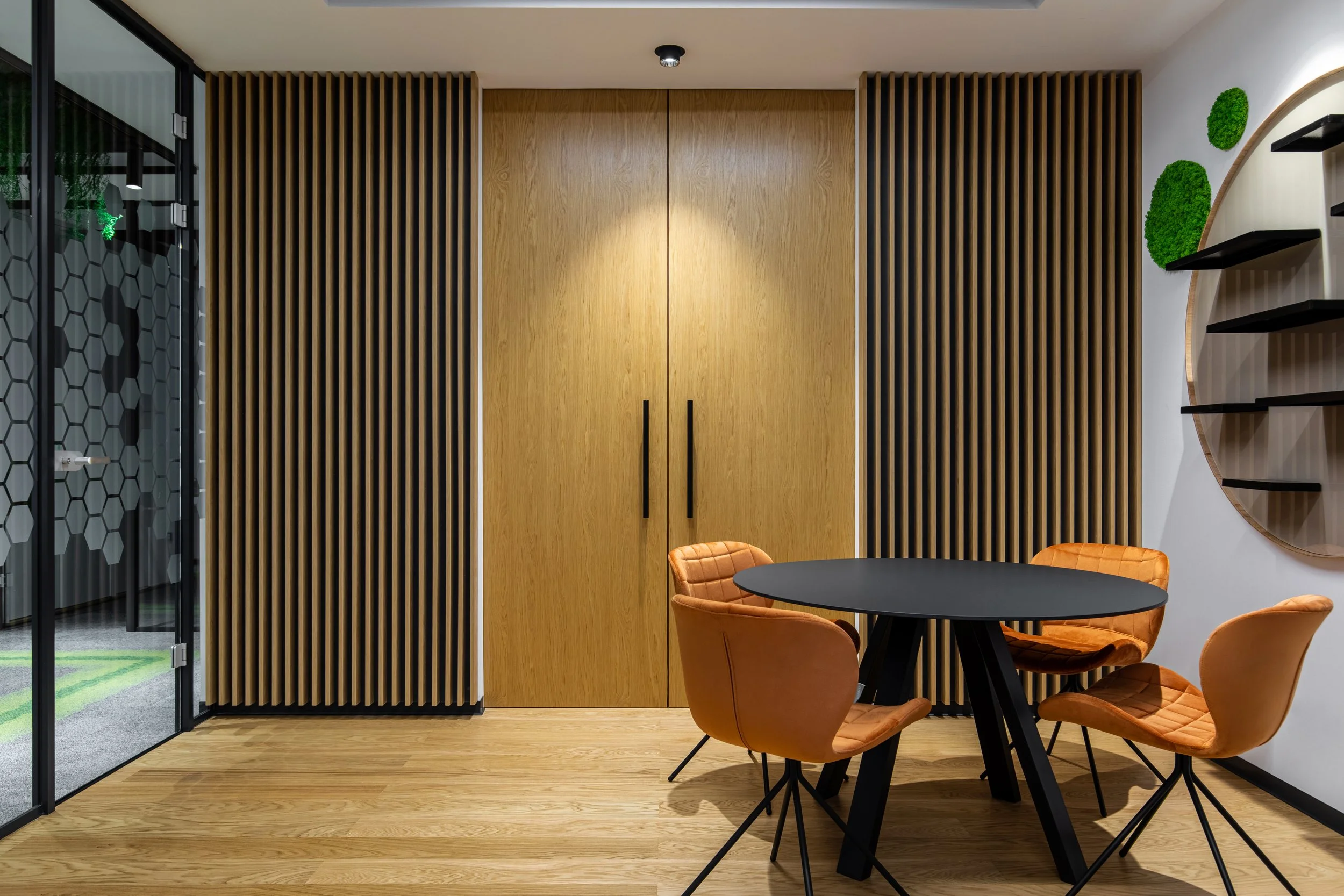 Modern interior room with a black round table and four orange upholstered chairs, wooden wall panels, and a glass door with a hexagonal pattern.