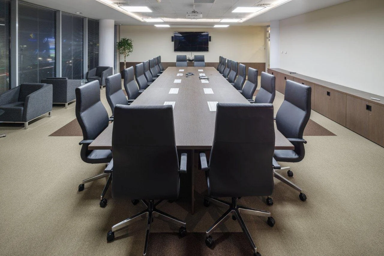 A modern conference room with a long rectangular table surrounded by black rolling office chairs, a TV mounted on the wall, and large windows with blinds on one side.