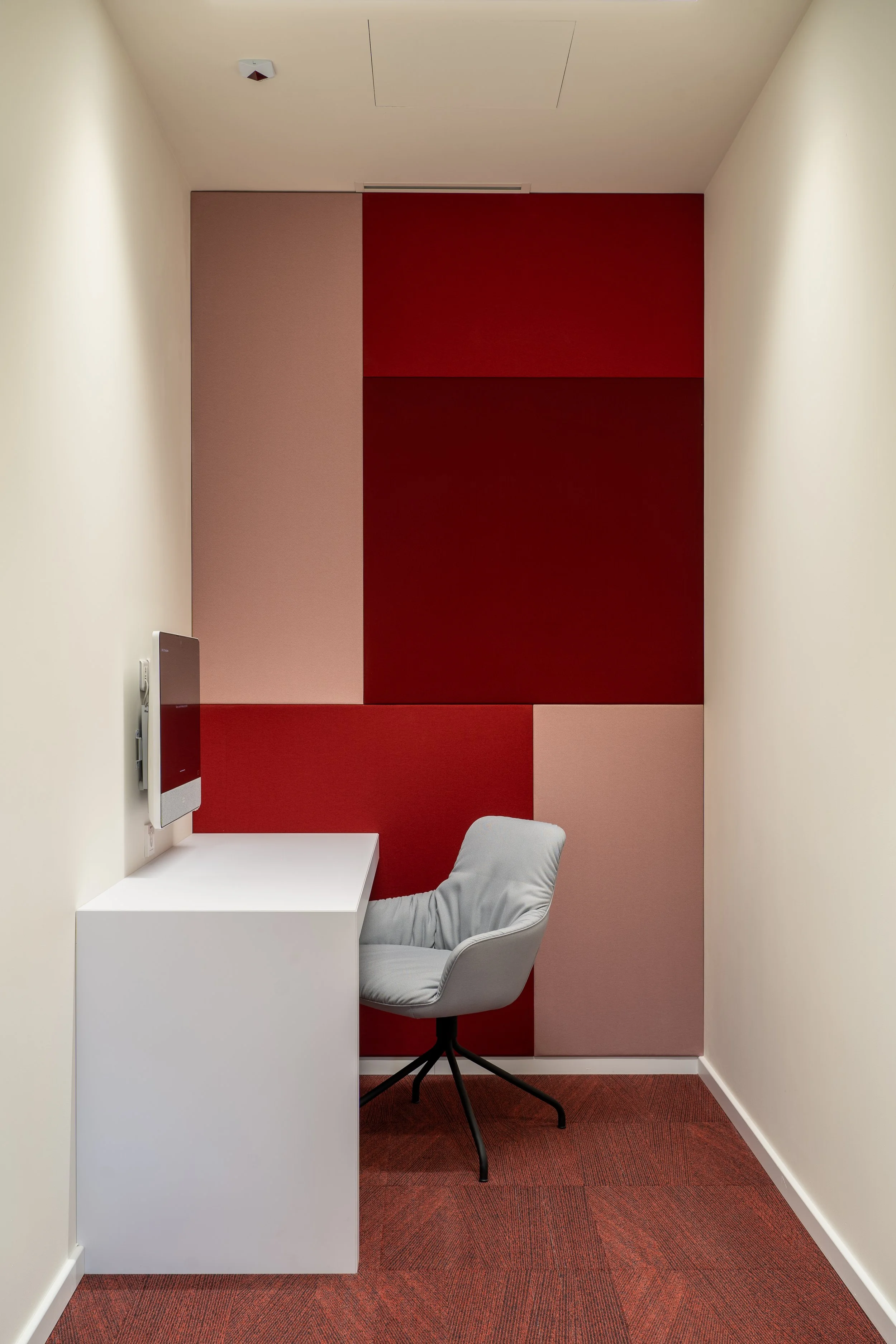 A small, private booth with a white desk, a comfortable gray chair, and a vibrant red and pink geometric acoustic wall panel.
