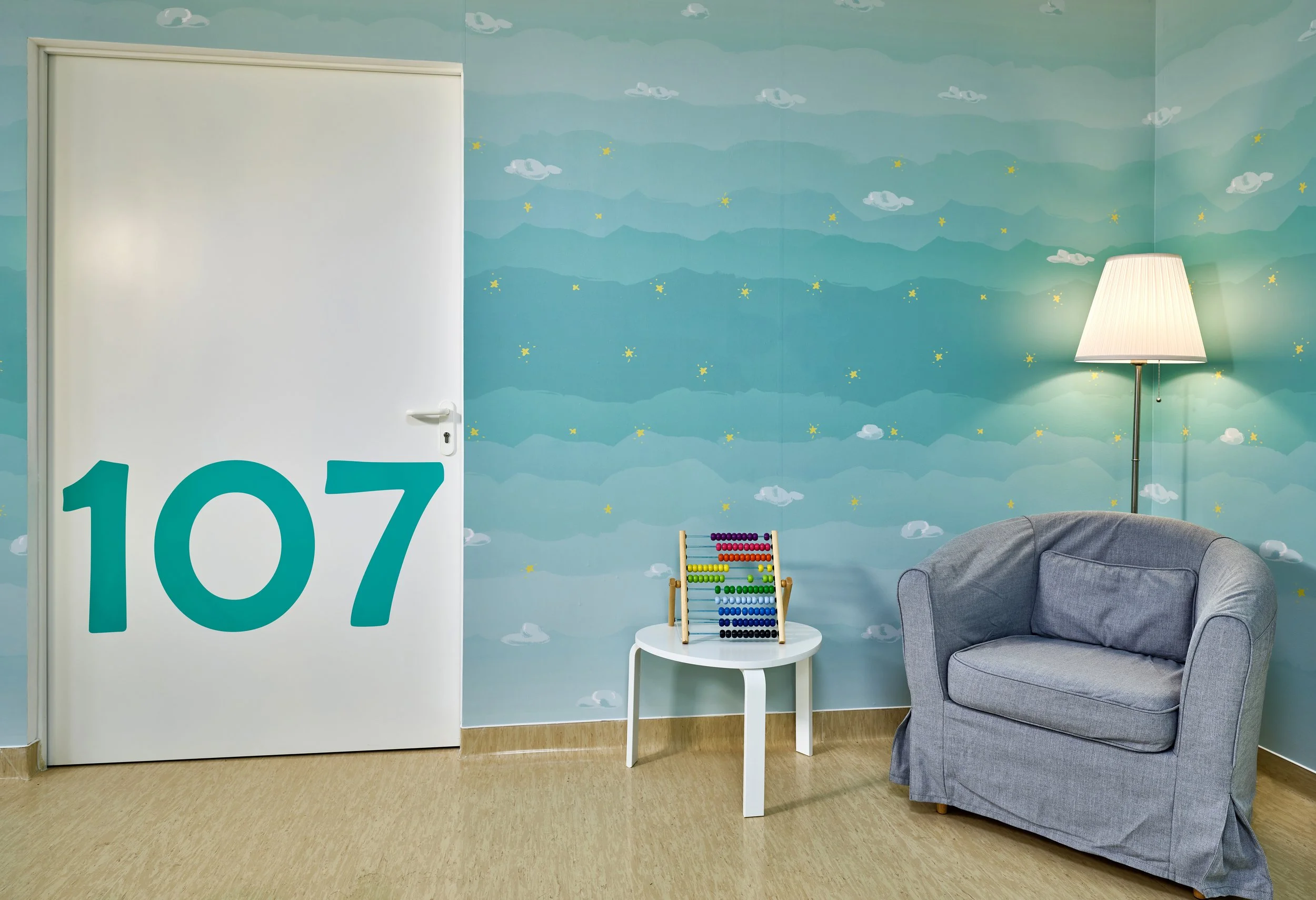 A room with a sky-themed mural on the wall, featuring clouds and stars, a white door with the number 107 on it, a small white table with a colorful abacus, a gray armchair, and a tall floor lamp with a white shade.