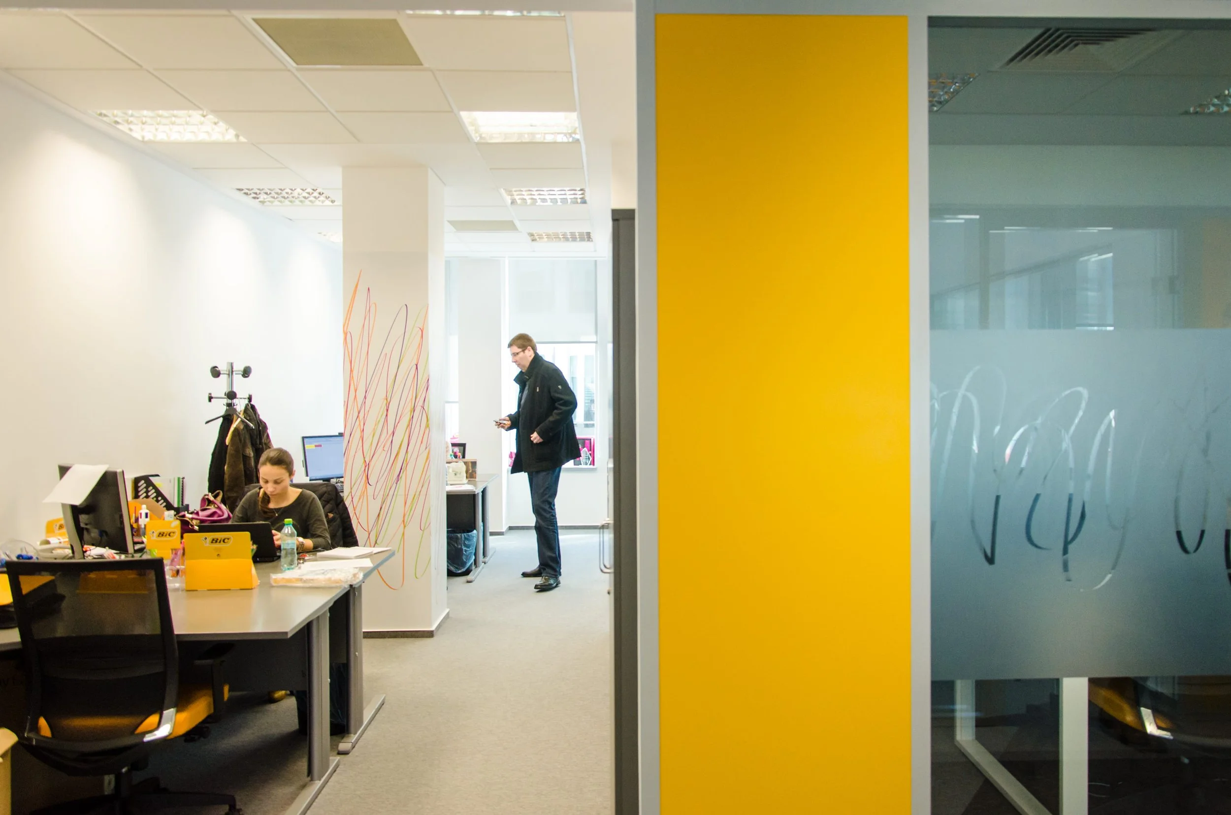An office workspace viewed through a glass wall with a yellow accent panel. Inside, a woman is working at a desk with a computer, and a man is standing in the background looking at his phone.