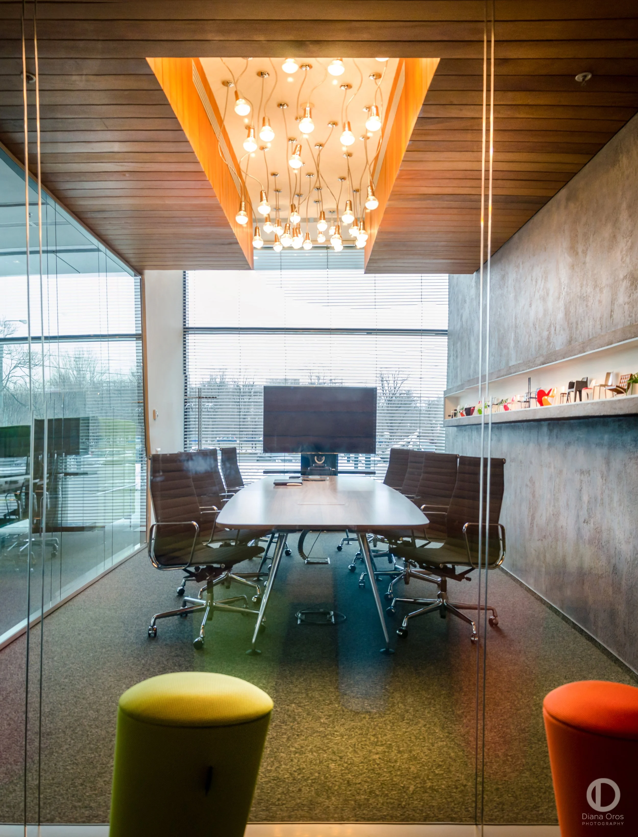 Modern conference room with a large wooden table surrounded by black office chairs, a flat screen monitor on the table, large windows with blinds, a colorful pendant light fixture hanging from a wood-paneled ceiling, and a glass wall with colorful stools outside.