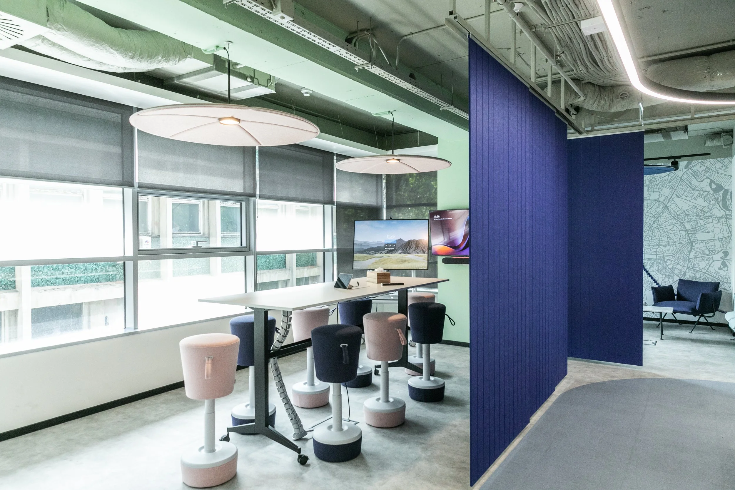 Modern office space with large windows, hanging circular lights, and a high table with colorful stools. Multiple monitors are mounted on a green wall, and there is a sitting area with a sofa and chairs in the background. A blue partition separates different areas of the office.