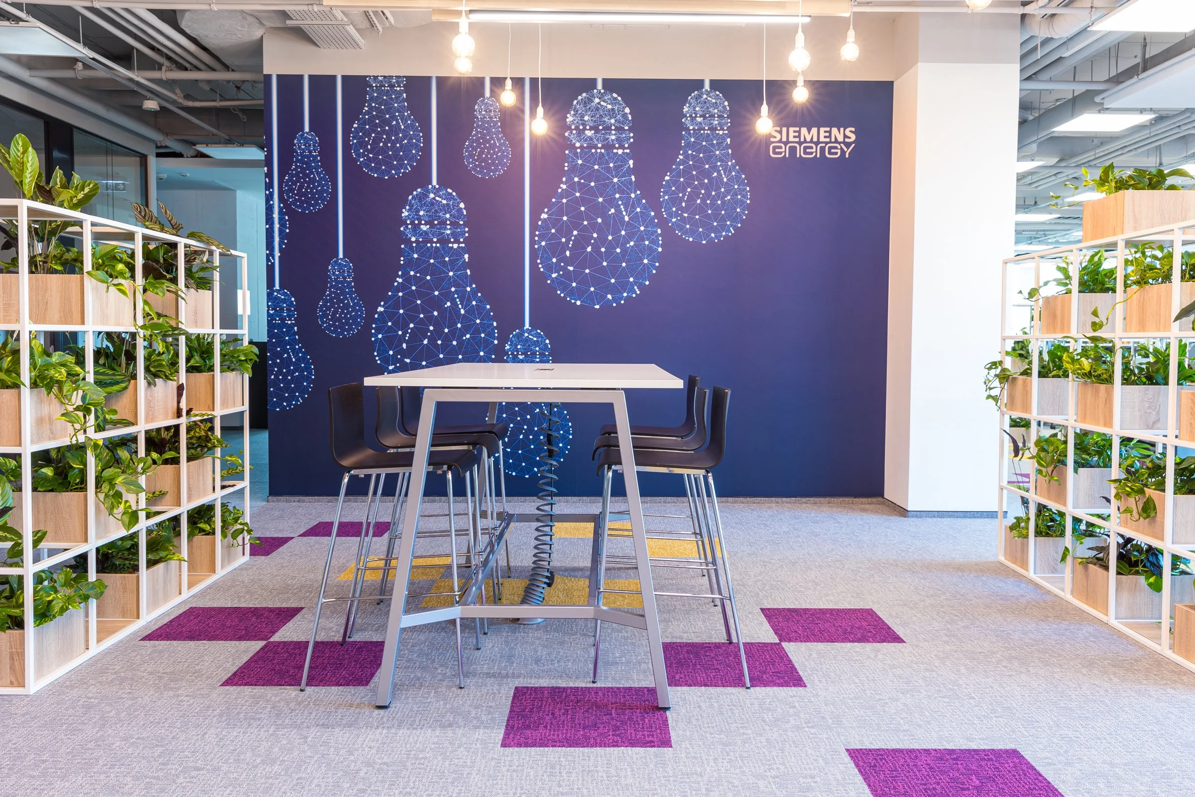Modern office space with a white high table and black chairs, framed by plant shelves on either side, in front of a blue wall with a pattern of illuminated hanging light bulb graphics and Siemens Energy logo.