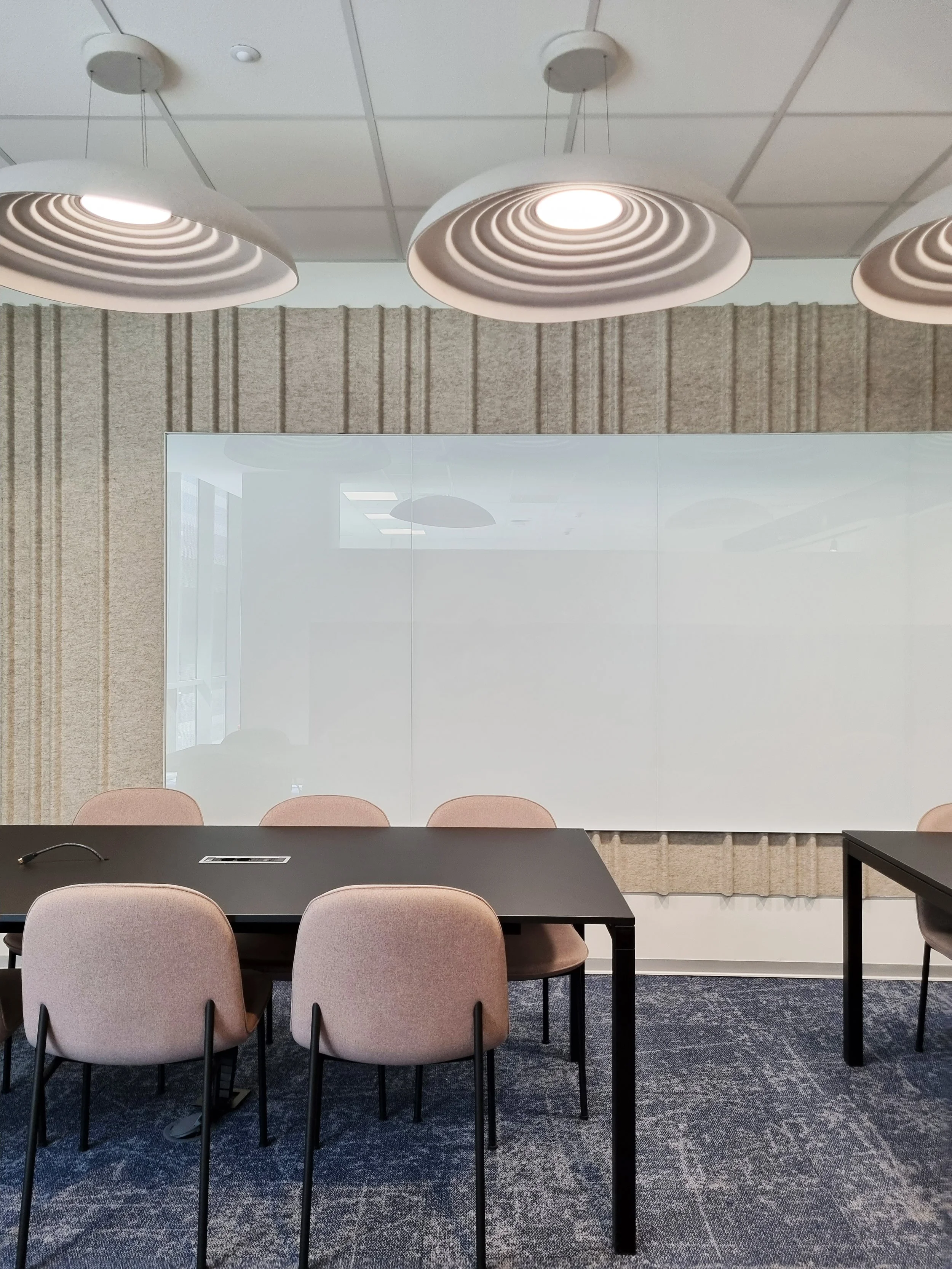 An empty conference room with a glass wall, a black table, beige chairs, overhead lighting fixtures, and a whiteboard.