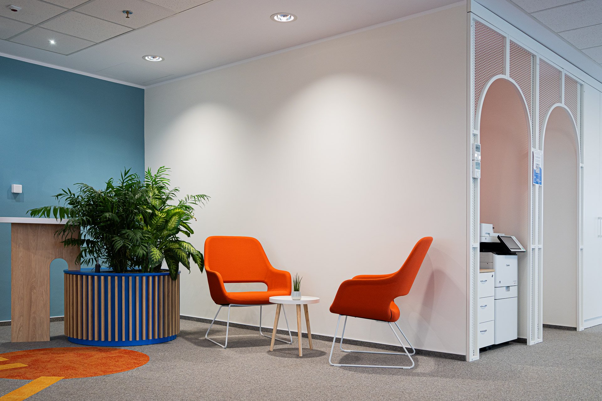 Office lobby with two orange chairs, a small round table with a potted plant, a large green plant in a modern blue planter, a gray carpet, white and blue walls, and a reception area with a copier.