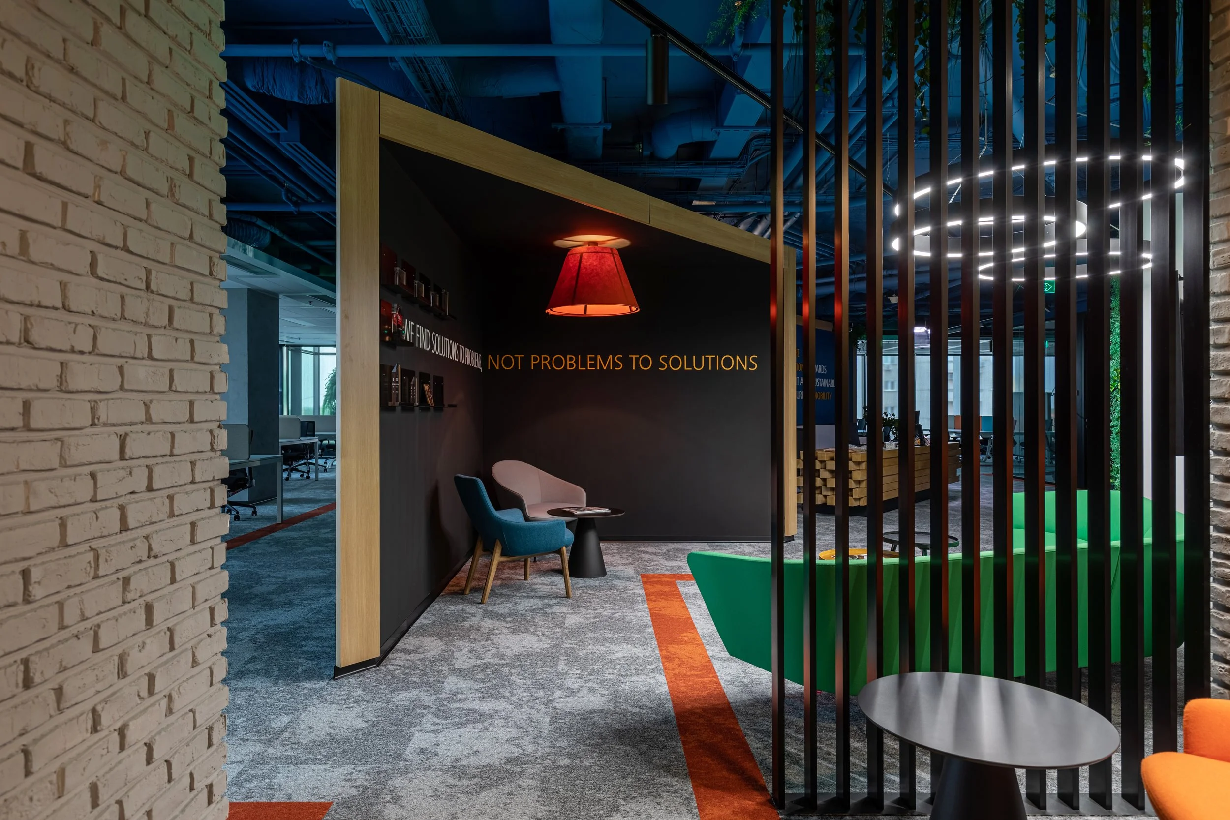 Modern office lounge area with seating, a black wall with the phrase 'NOT PROBLEMS TO SOLUTIONS', and colorful decor including green and orange furniture, enclosed partly by black vertical slats.