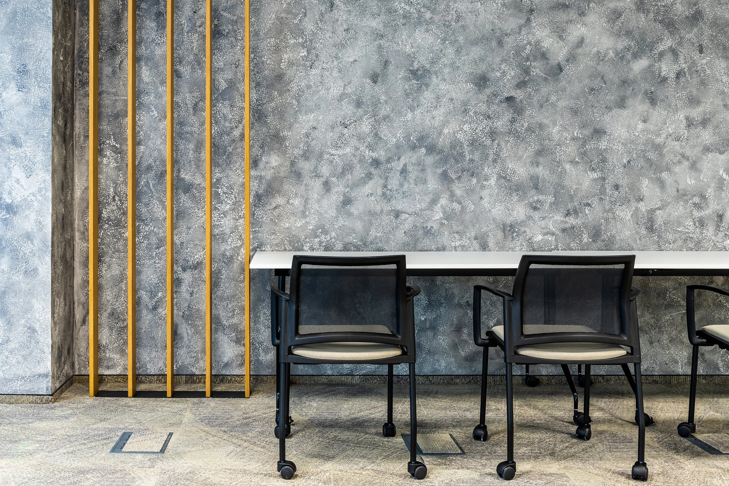 Modern office meeting space with three black mesh chairs with beige cushions, a white table, a textured grey wall, and gold vertical slats on the side.