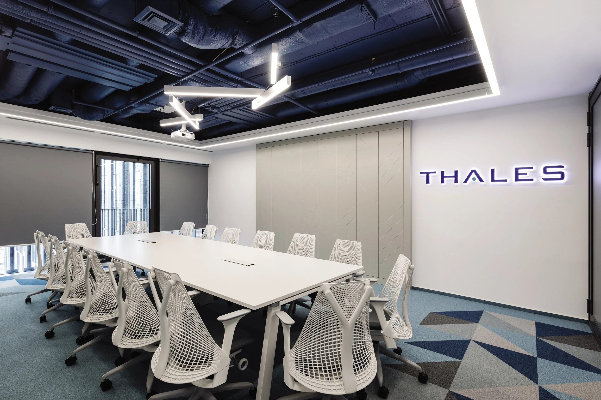 Modern conference room with a long white table, white ergonomic chairs, a ceiling with dark exposed pipes, LED strip lighting, large windows with gray blinds, a geometric carpet, and a wall with the illuminated Thales logo.
