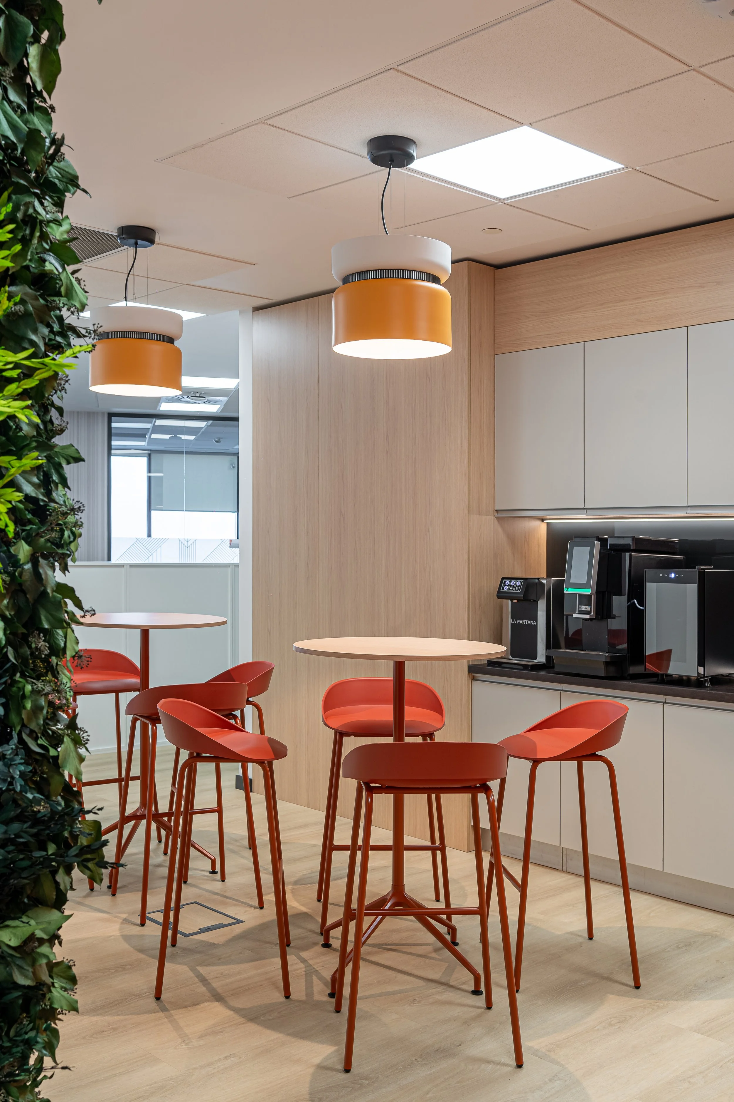 Modern break room or kitchenette area with two round pendant lights, four red chairs around two small round tables, a wall with built-in cabinets, and a coffee machine.