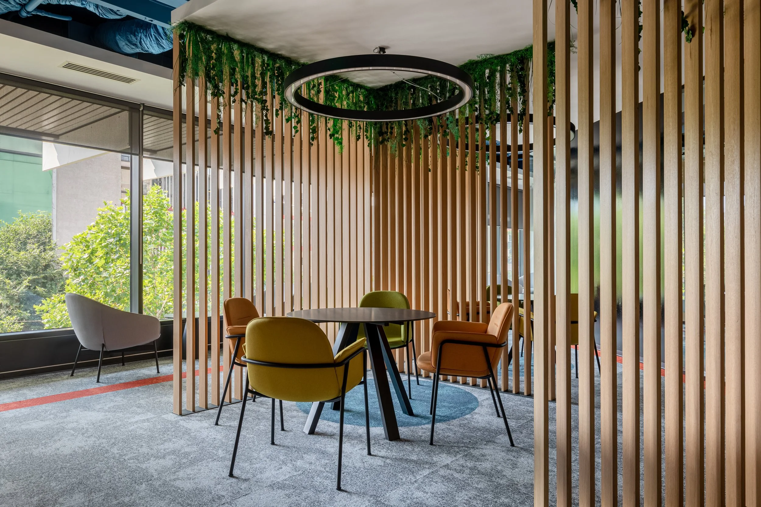 Interior of a modern office meeting room with wooden slat walls, a round black table, colorful chairs, a circular ceiling light, and large windows with a view of trees outside.