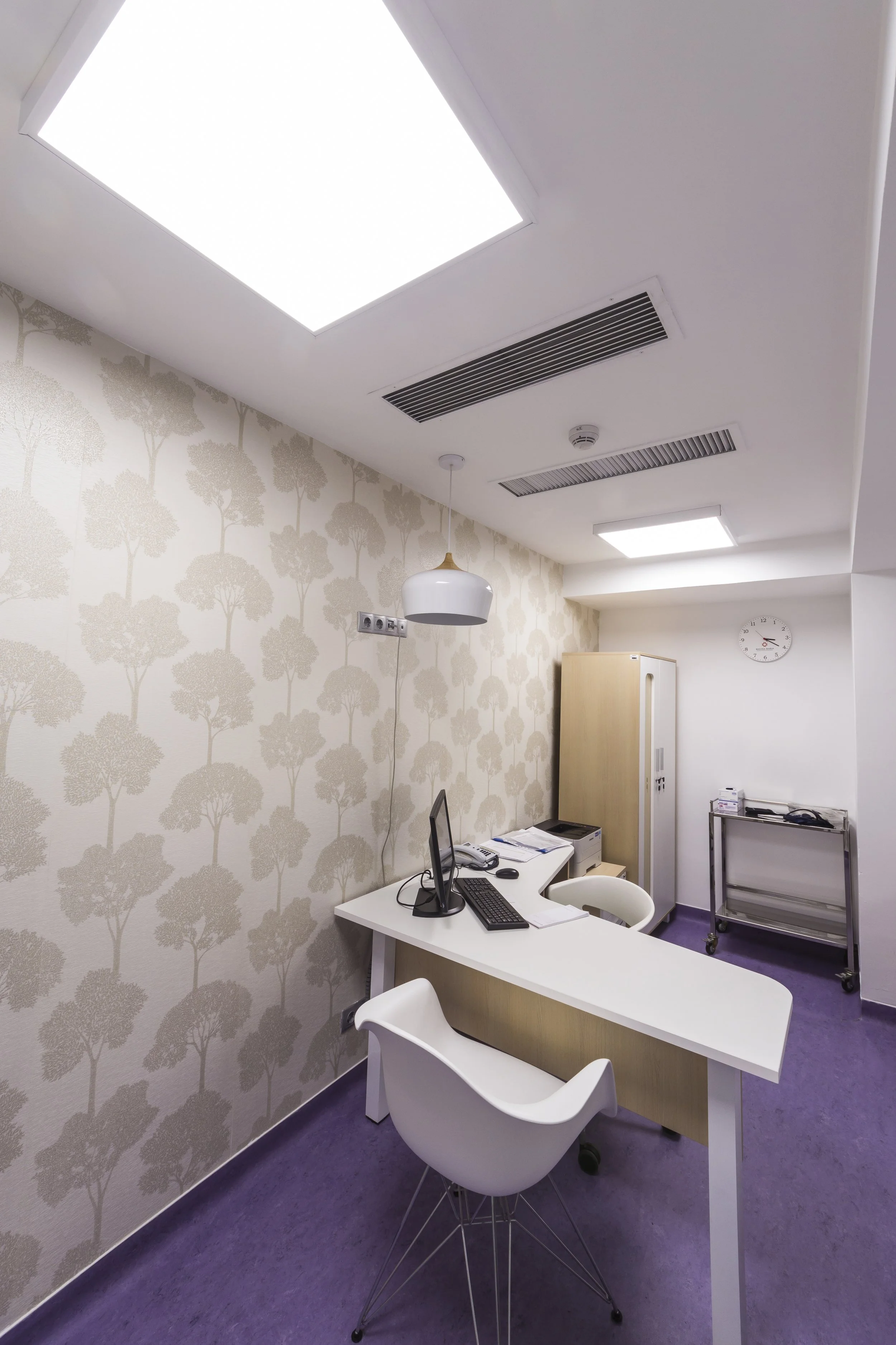 Empty office with white desk, two white chairs, computer, telephone, printer, and file cabinet, purple carpet, patterned wallpaper, ceiling lights, clock on white wall, and a cart.