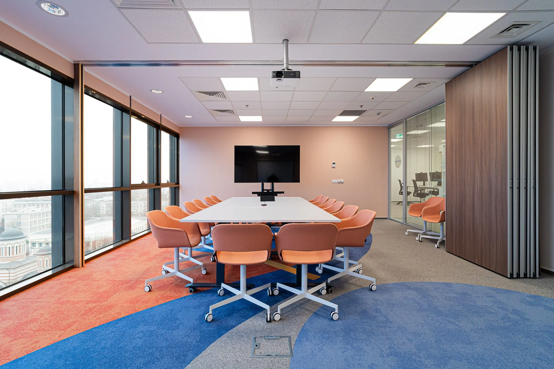 Empty modern conference room with large windows, a long white table surrounded by peach-colored chairs, a wall-mounted TV, and a glass-walled office space to the side.