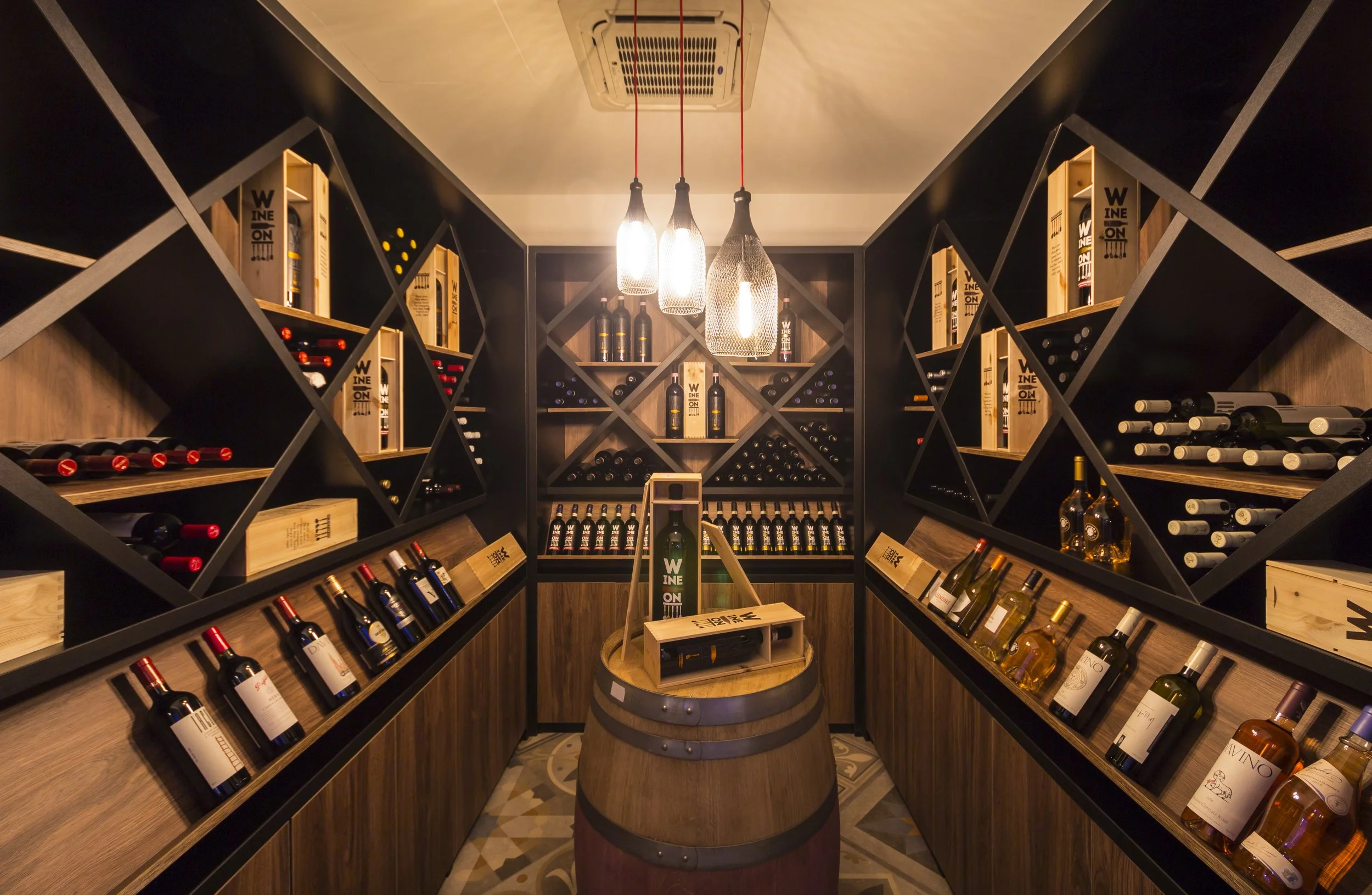 A wine cellar with wooden shelves filled with bottles of wine on both sides, a barrel in the center with a small display, and modern hanging lights overhead.