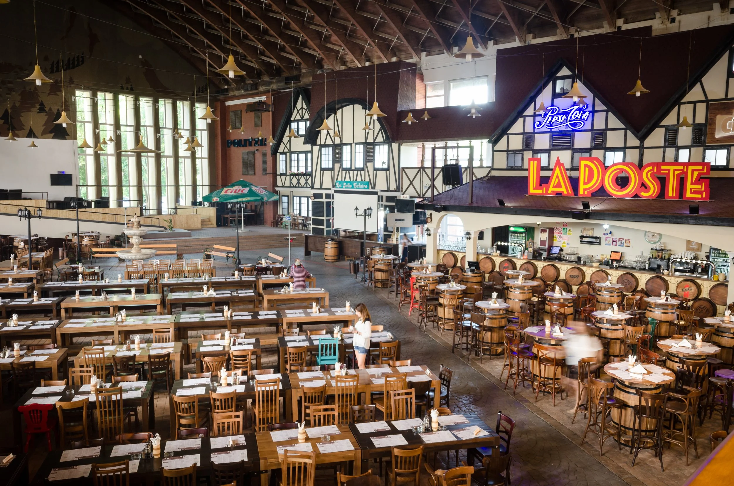 Inside a large restaurant with many wooden tables and chairs, some set with menus and silverware. A bar area with barrels and stools is visible on the right, with bright, colorful signs including 'La Poste' and neon signage. Large windows let in natural light, and there is a raised stage area with a small outdoor seating section in the background.