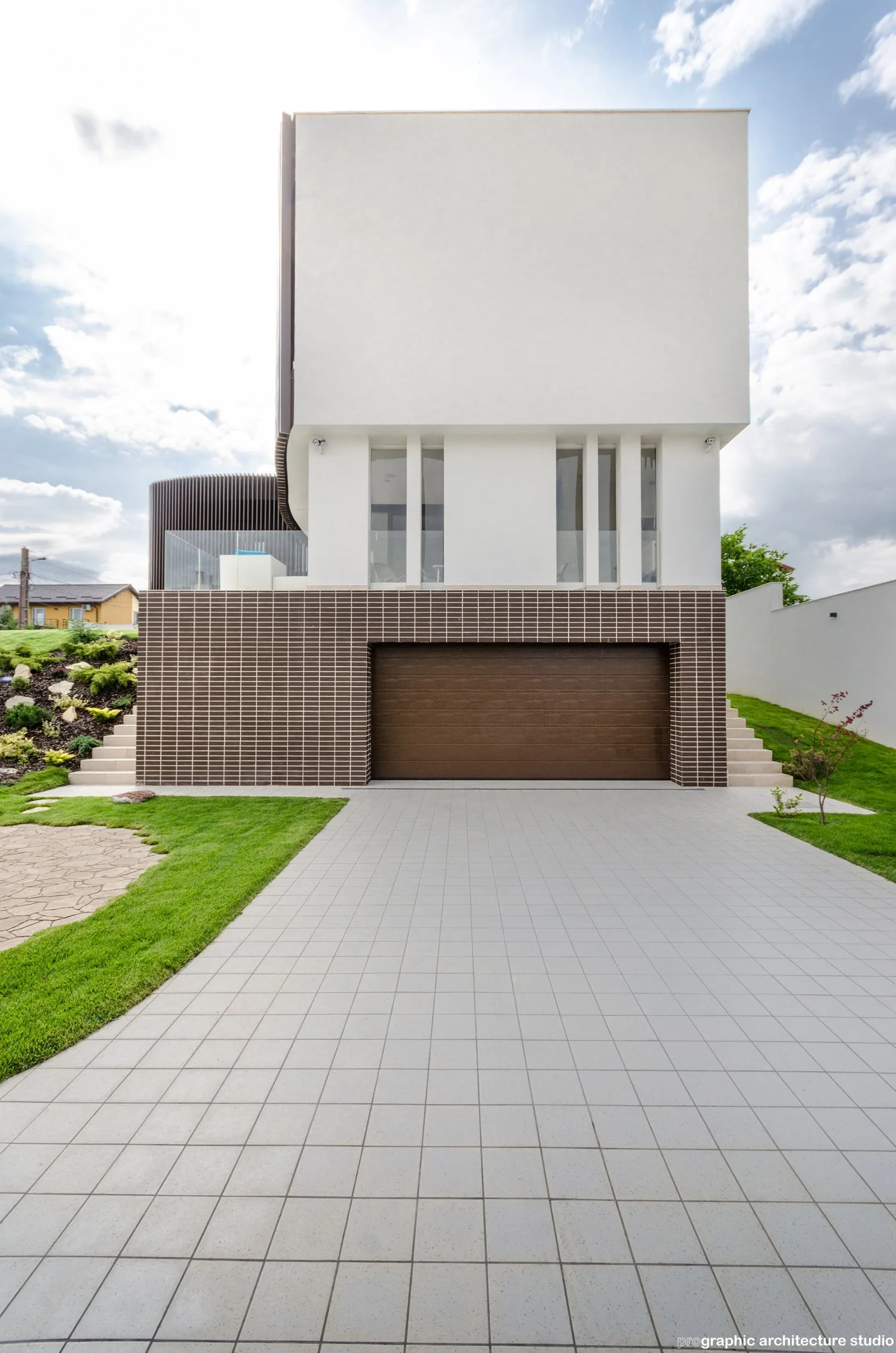 Front view of a modern white house with a brown garage door, stairs on either side, a tiled driveway, and landscaped lawn.