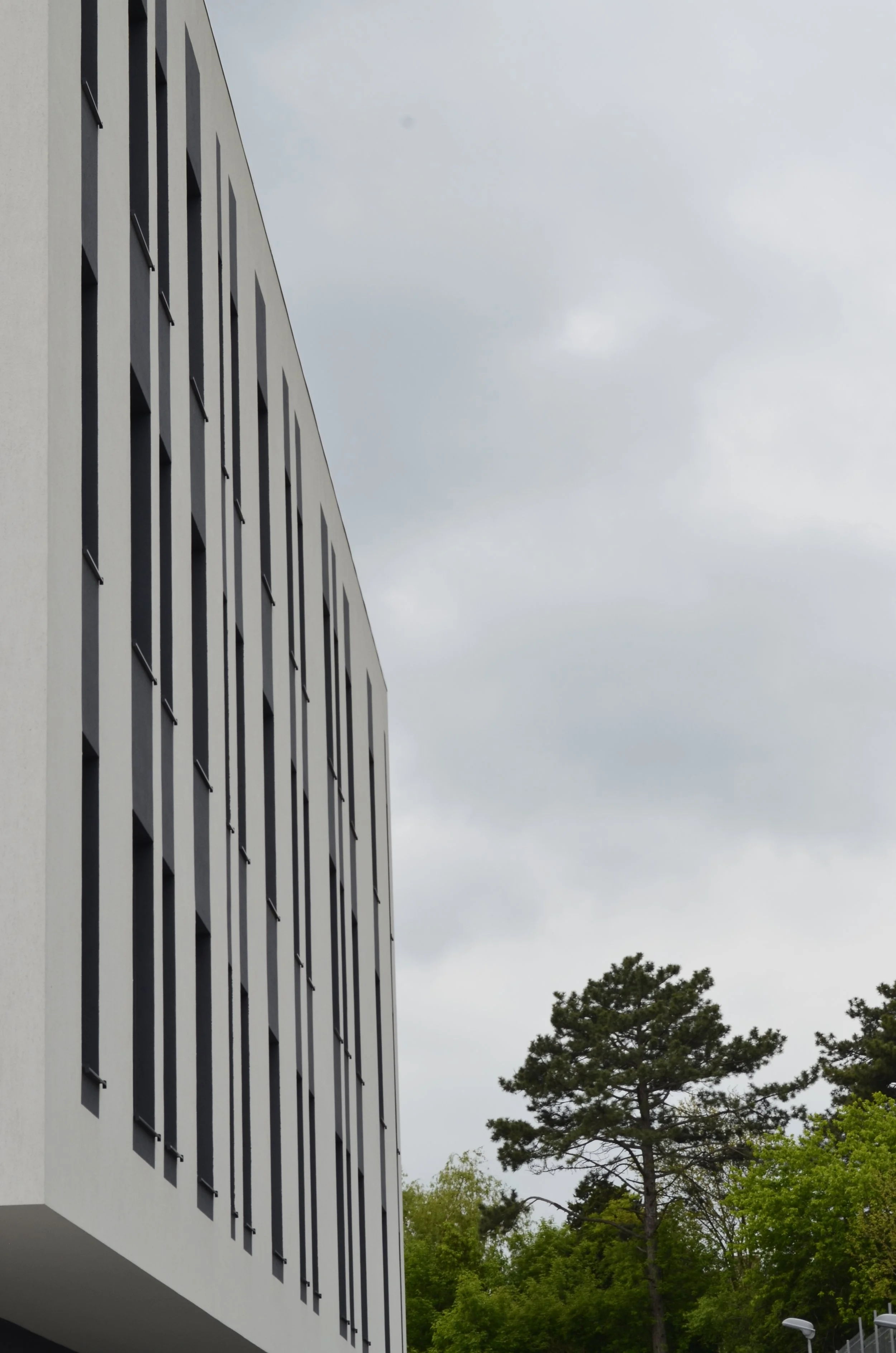 Modern white building with vertical black window slats, green trees, and a cloudy sky in the background.