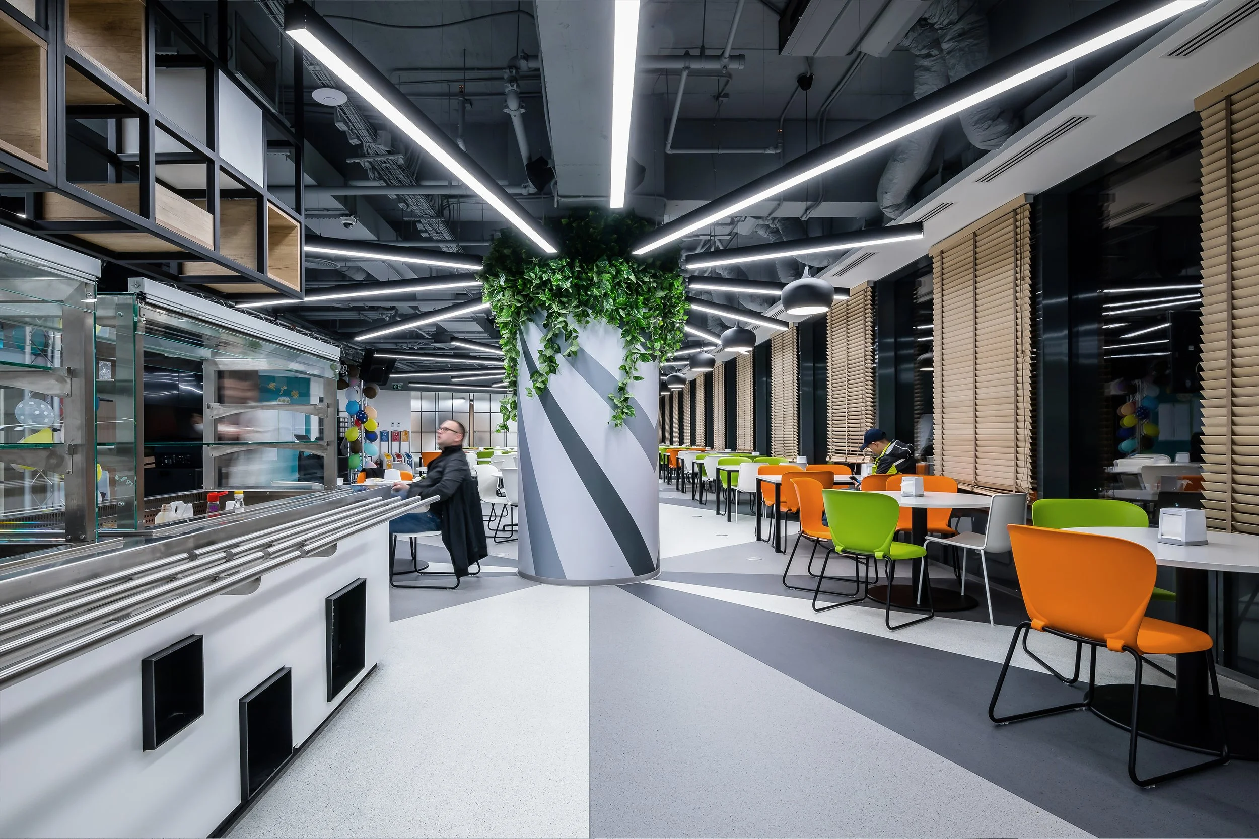 Modern cafeteria with colorful chairs, large central column with greenery, and black ceiling with exposed ductwork and linear lighting.