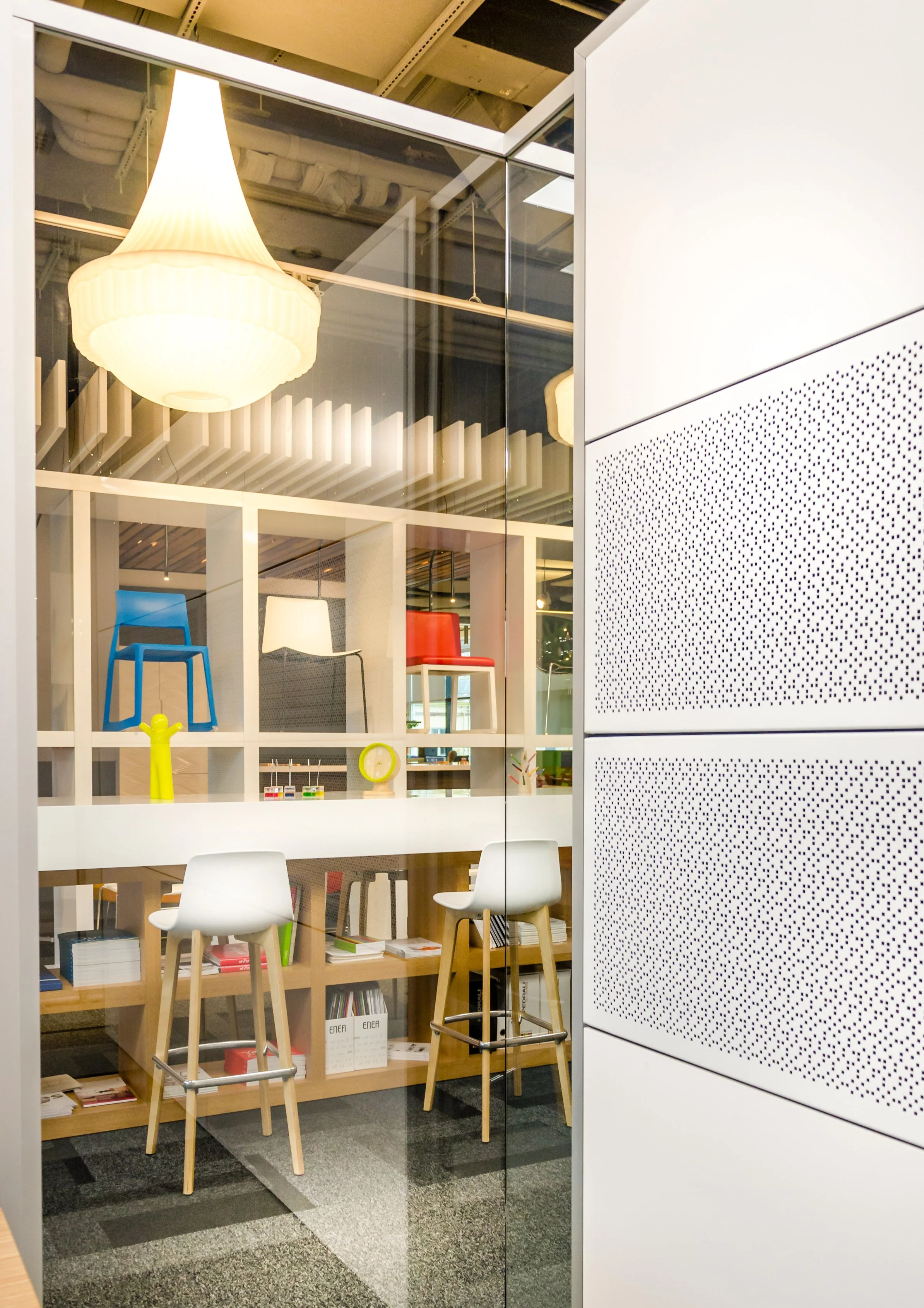 Interior view of a modern office space showing a glass wall, white chairs with wooden legs, and colorful chairs displayed on a wall shelf.
