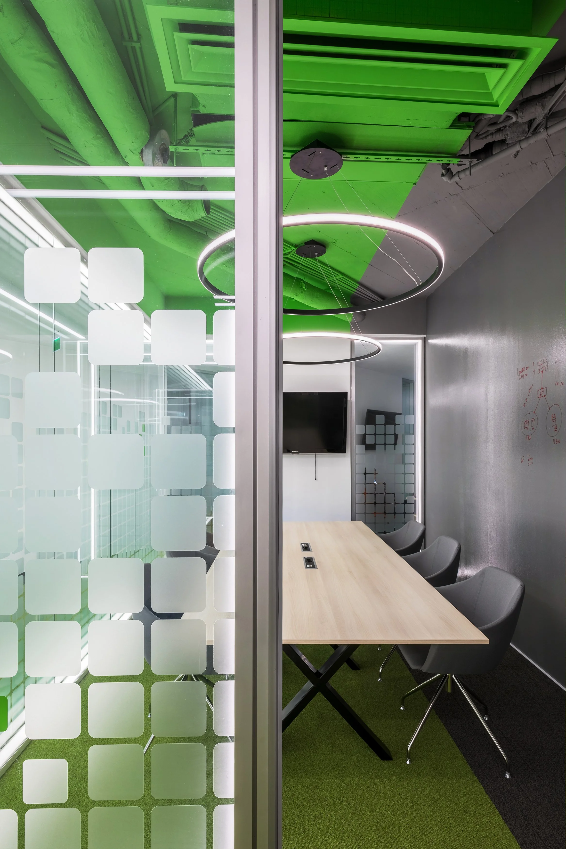 Modern office meeting room with a wooden table, four gray chairs, a flat-screen TV, and contemporary circular LED pendant lights. The room has gray walls and a partial glass wall with a frosted square pattern separating it from the rest of the office. The ceiling is painted green with visible pipes and ductwork.