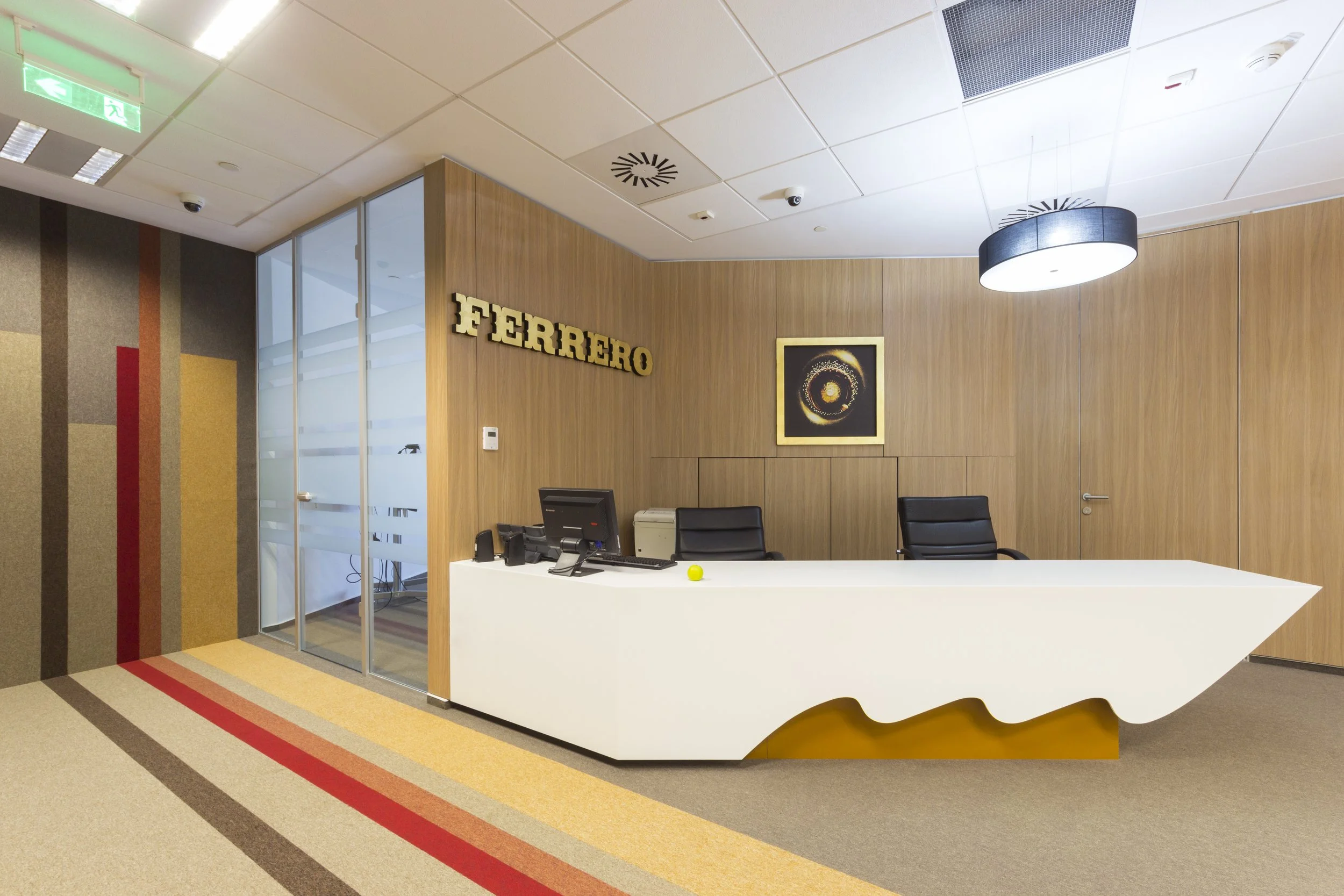 FERRERO reception area with a white modern desk, black chairs, a computer, a framed art piece on wood-paneled wall, and the FERRERO logo in gold letters, with multicolored striped carpet and glass door on the left.