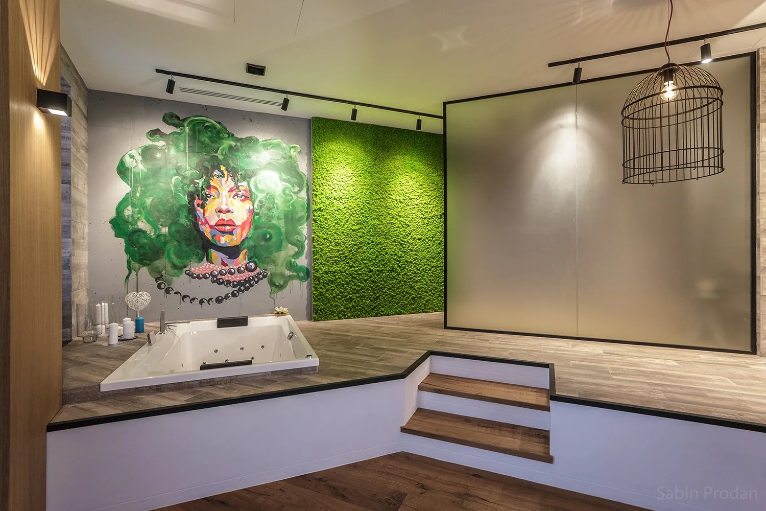 Interior of a modern bathroom or spa with a small white jacuzzi tub, colorful graffiti portrait on the wall, green moss wall, and a large frosted glass partition. Decorative candles and jars are on the ledge.
