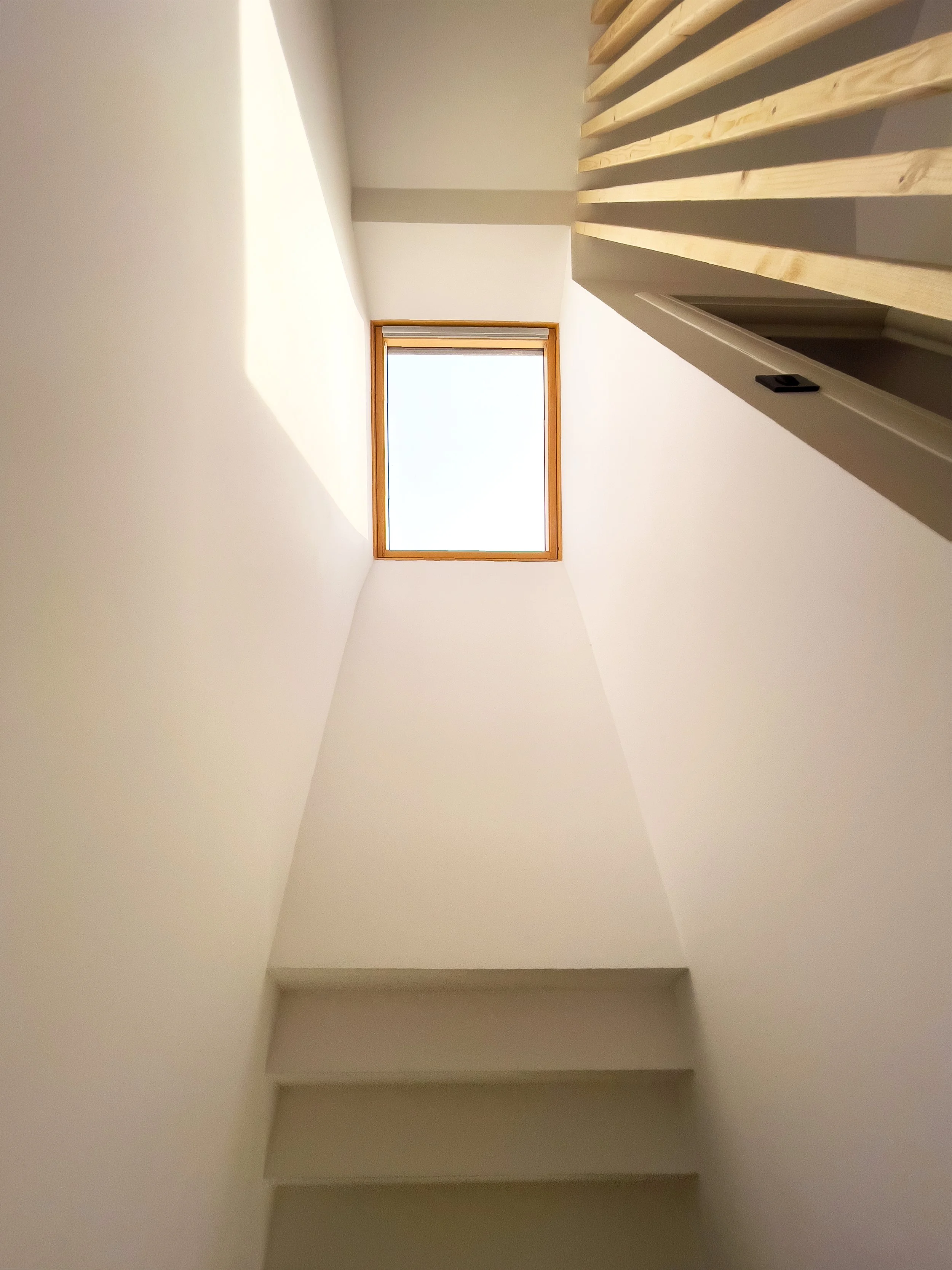 Looking upward at a stairwell with white walls, four steps, and a square window at the top letting in natural light.