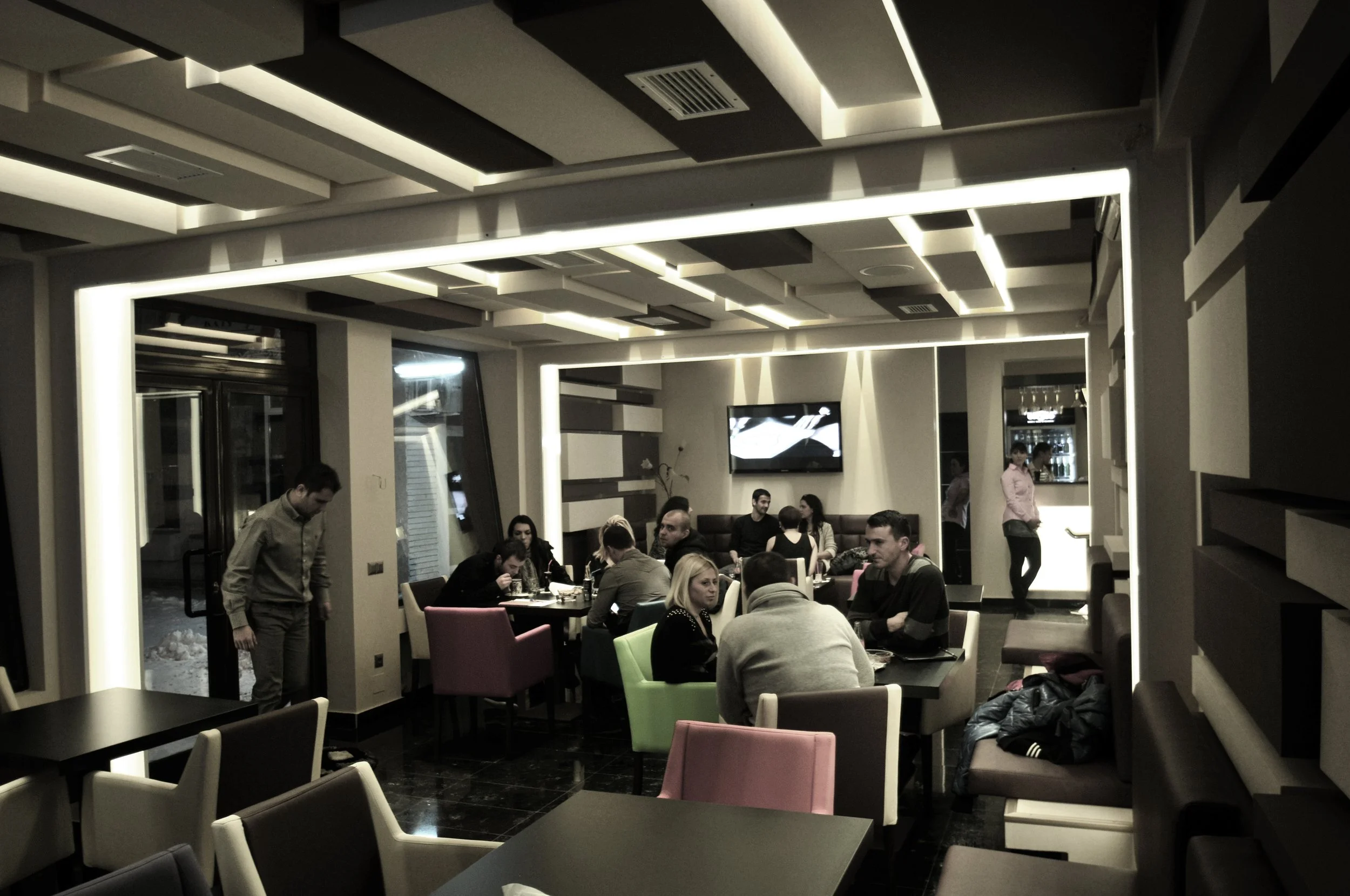 Interior of a modern restaurant or cafe with hanging ceiling lights, a TV screen on the wall, and several people sitting at tables talking and eating.