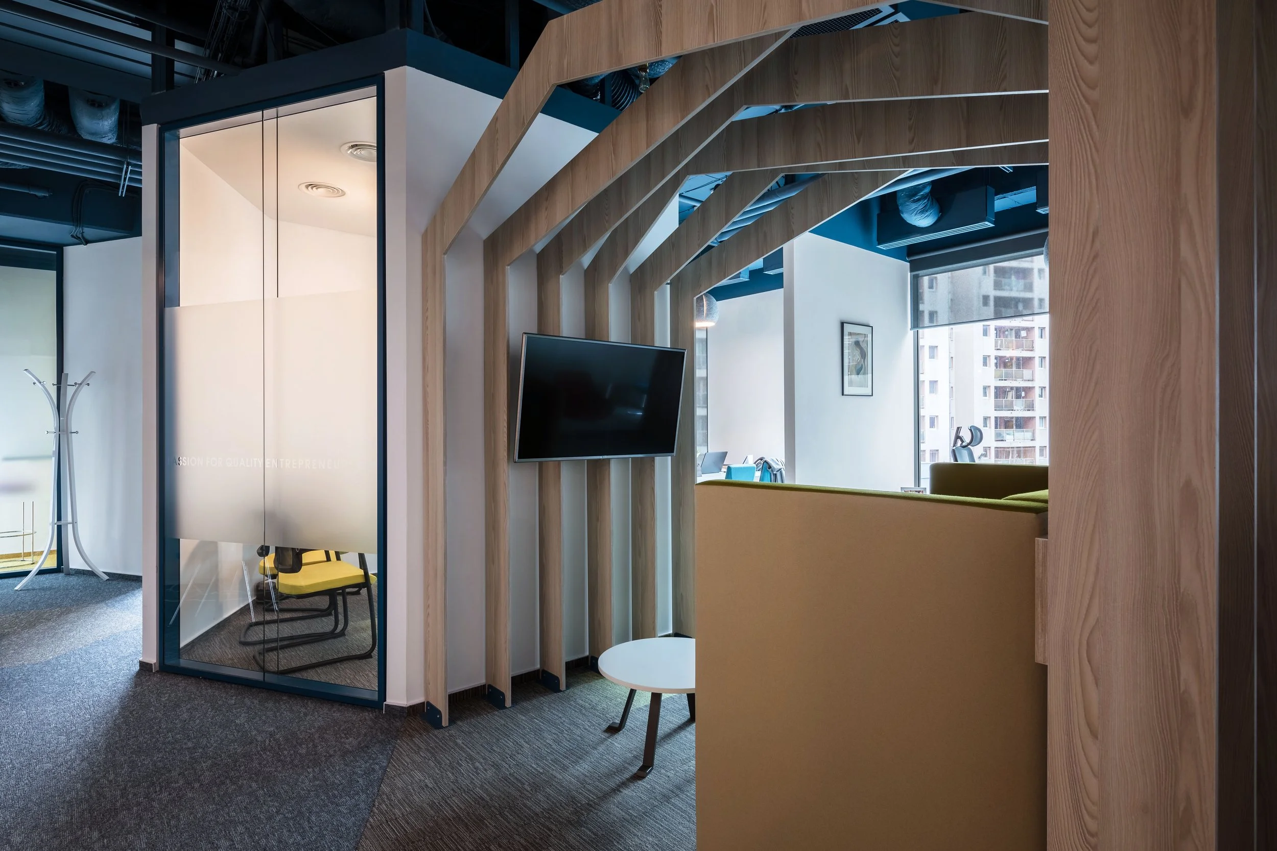 Modern office space with wooden architectural features, a mounted television, a glass-walled meeting room with a yellow chair inside, and city buildings visible through the windows.