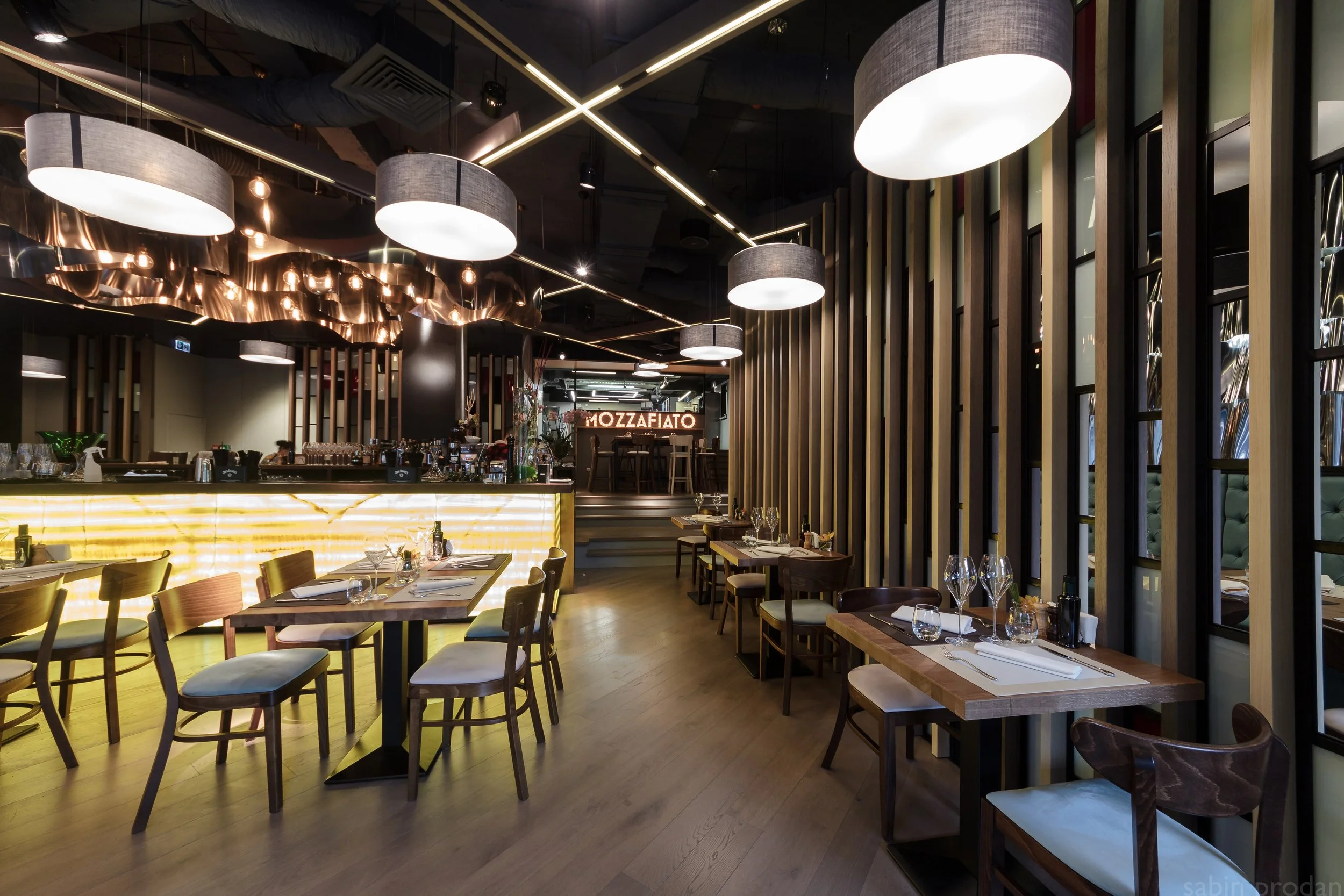 Interior view of a modern restaurant with wooden furniture, hanging lights, and a bar area in the background. The restaurant appears elegant with neatly set tables, wine glasses, and decorative lighting.