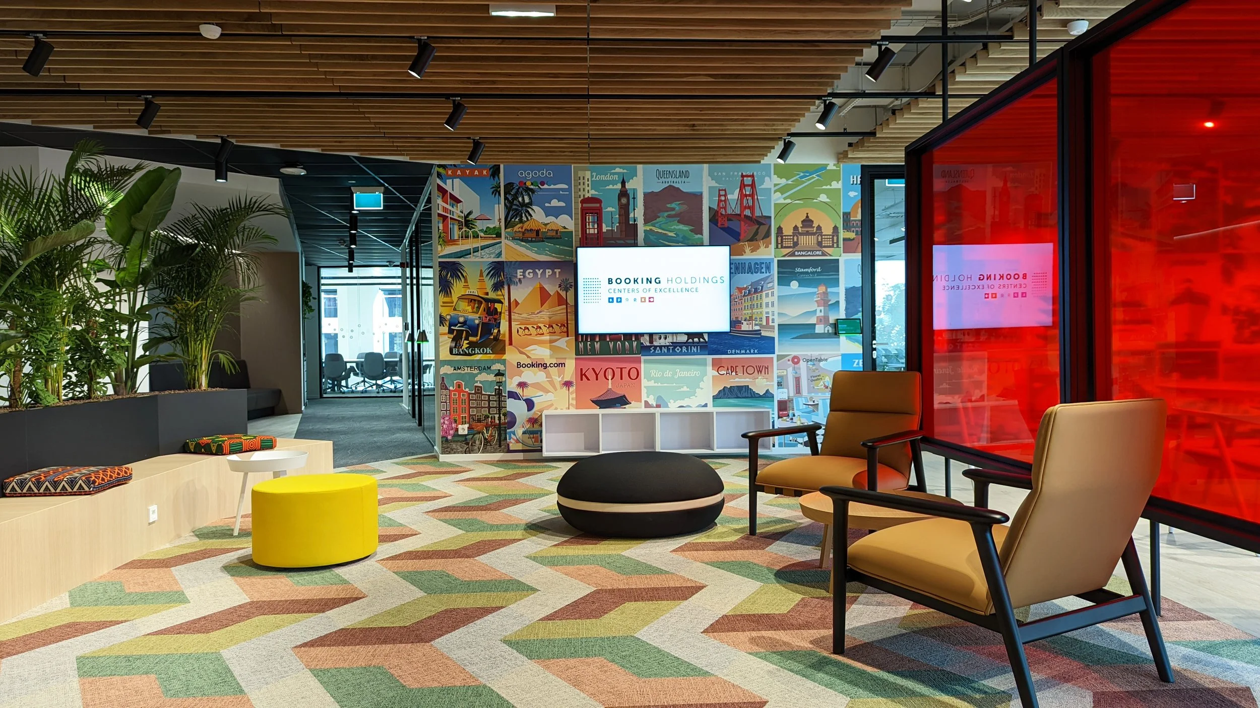 Modern office lounge with seating, green plants, colorful patterned carpet, and a wall with travel posters and a digital screen displaying 'Booking Holdings Centers of Excellence'