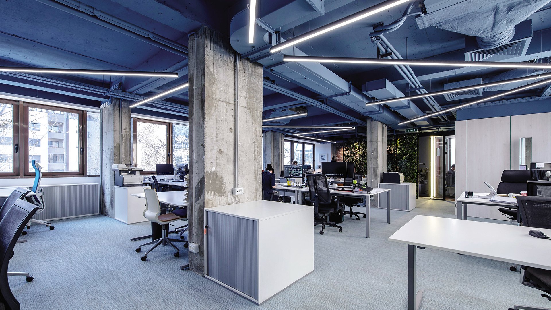 Modern office with concrete pillars, large windows, and suspended linear LED lights, featuring desks, chairs, and computers.