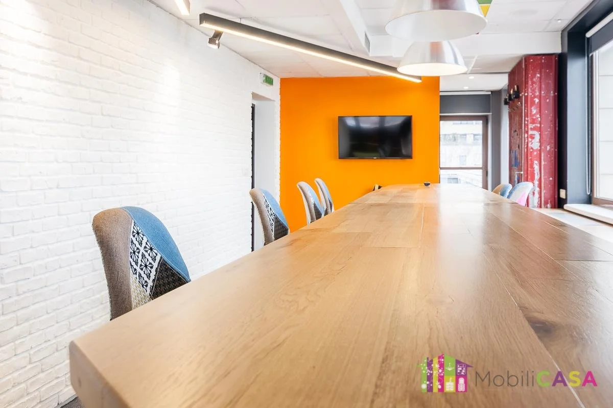 Modern conference room with a long wooden table, upholstered chairs, a white brick wall, a bright orange accent wall with a flat-screen TV, and large windows letting in natural light.