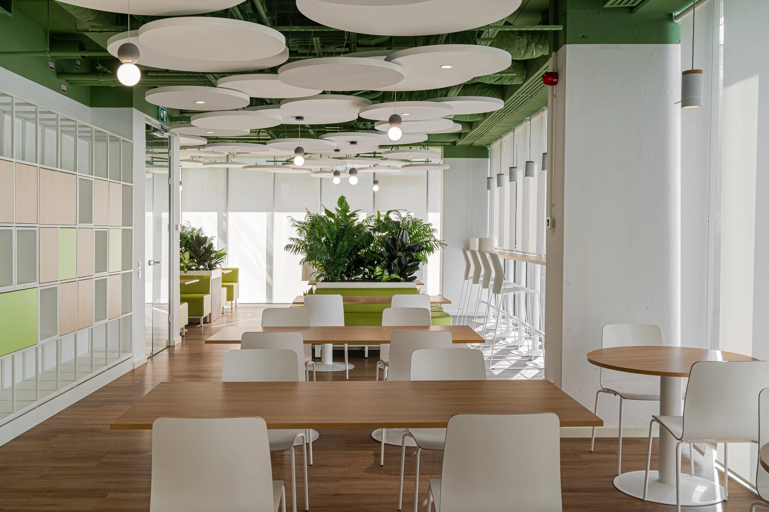 Modern indoor lounge or cafeteria with wooden tables and white chairs, large green plants, and decorative white ceiling panels with hanging lights.