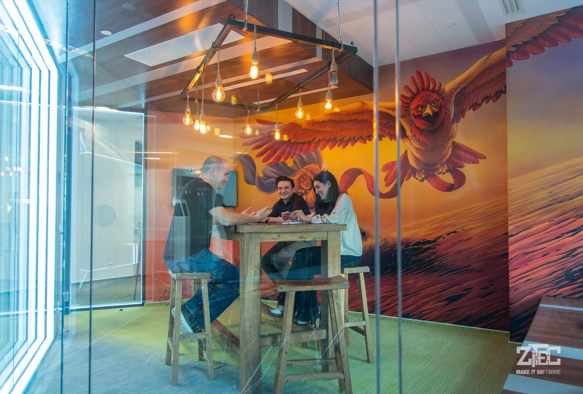 Three people sitting at a wooden high table inside a colorful room with a large mural of a red and orange eagle flying over a sunset.